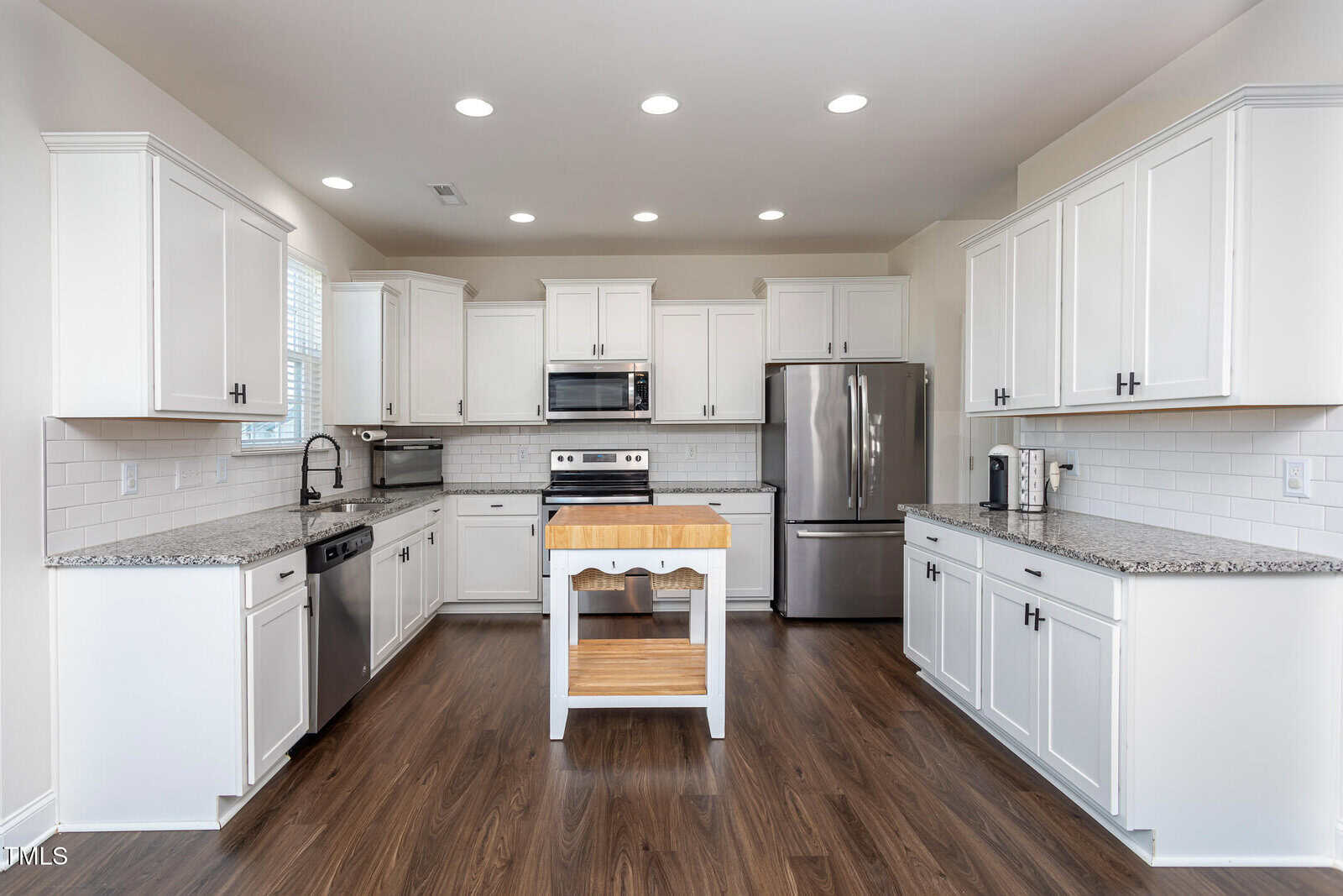 A modern kitchen with white cabinets, stainless steel appliances, a small wooden island in the center, granite countertops, subway tile backsplash, and dark wood flooring. Ceiling lights are on, creating bright illumination.