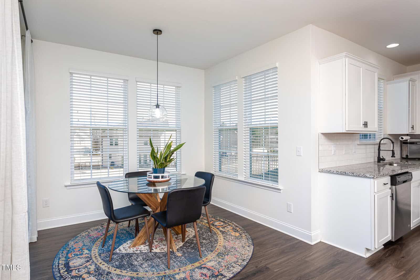 A modern dining area with a round glass table, four black chairs, a potted plant centerpiece, and a multicolored rug. Large windows with blinds let in natural light. White kitchen cabinets and granite countertops are visible.