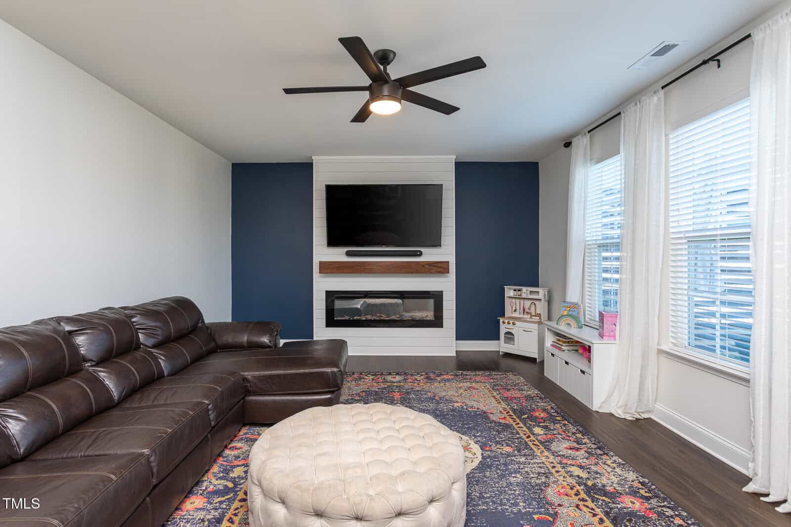 A living room with a dark brown leather sectional sofa, a tufted round ottoman, a patterned rug, a wall-mounted TV above a modern fireplace, a play kitchen set, and large windows with white curtains.