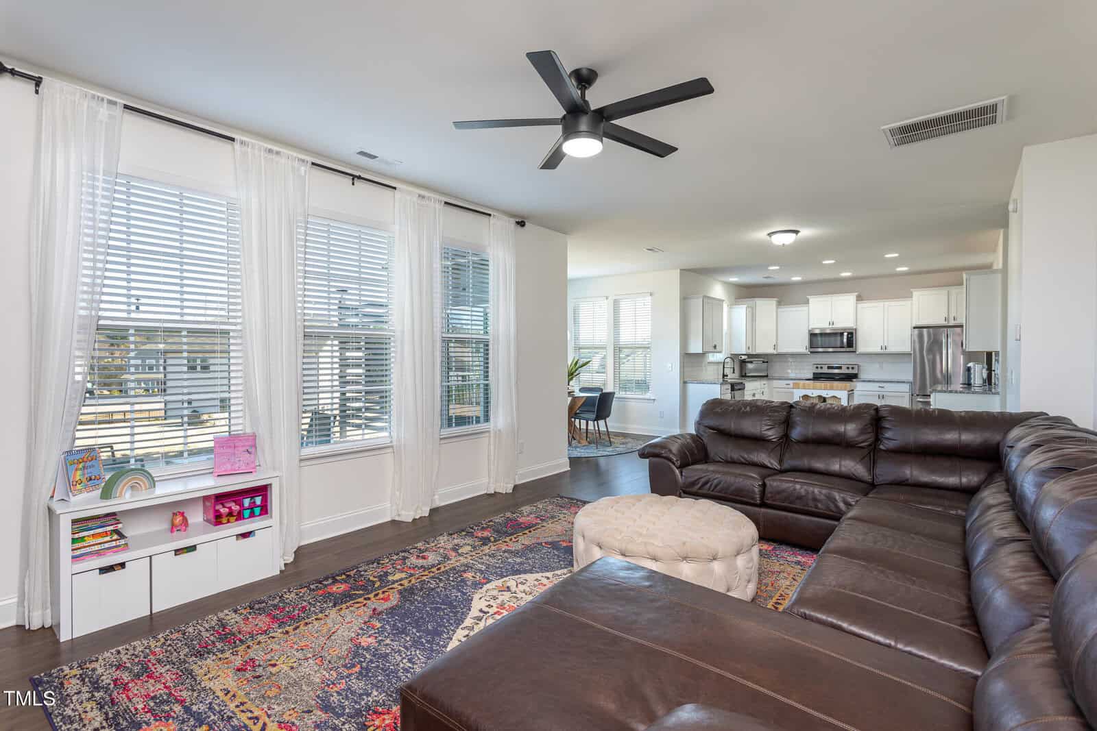 Living room with a large brown leather sectional, round ottoman, colorful rug, and built-in white shelves under three tall windows; kitchen and dining area are visible in the background. Ceiling fan and recessed lighting overhead.