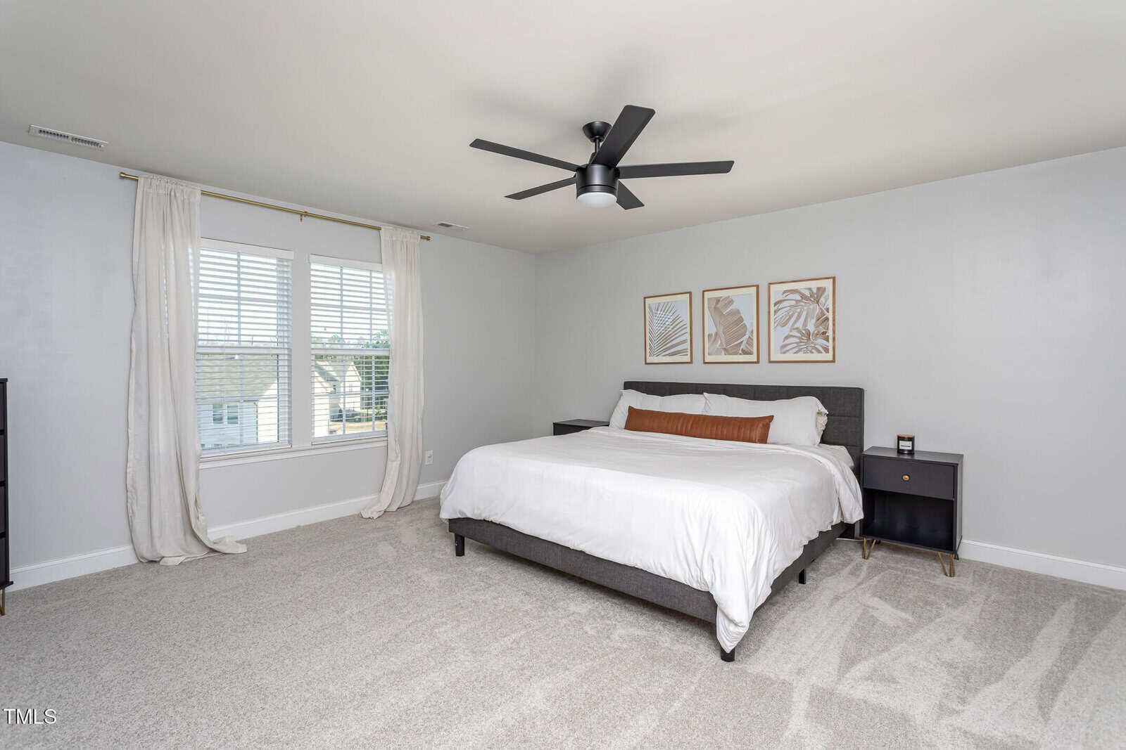 A bedroom with light walls and carpet, a large bed with white linens, two black nightstands, three framed prints above the bed, a black ceiling fan, and a window with white curtains letting in natural light.