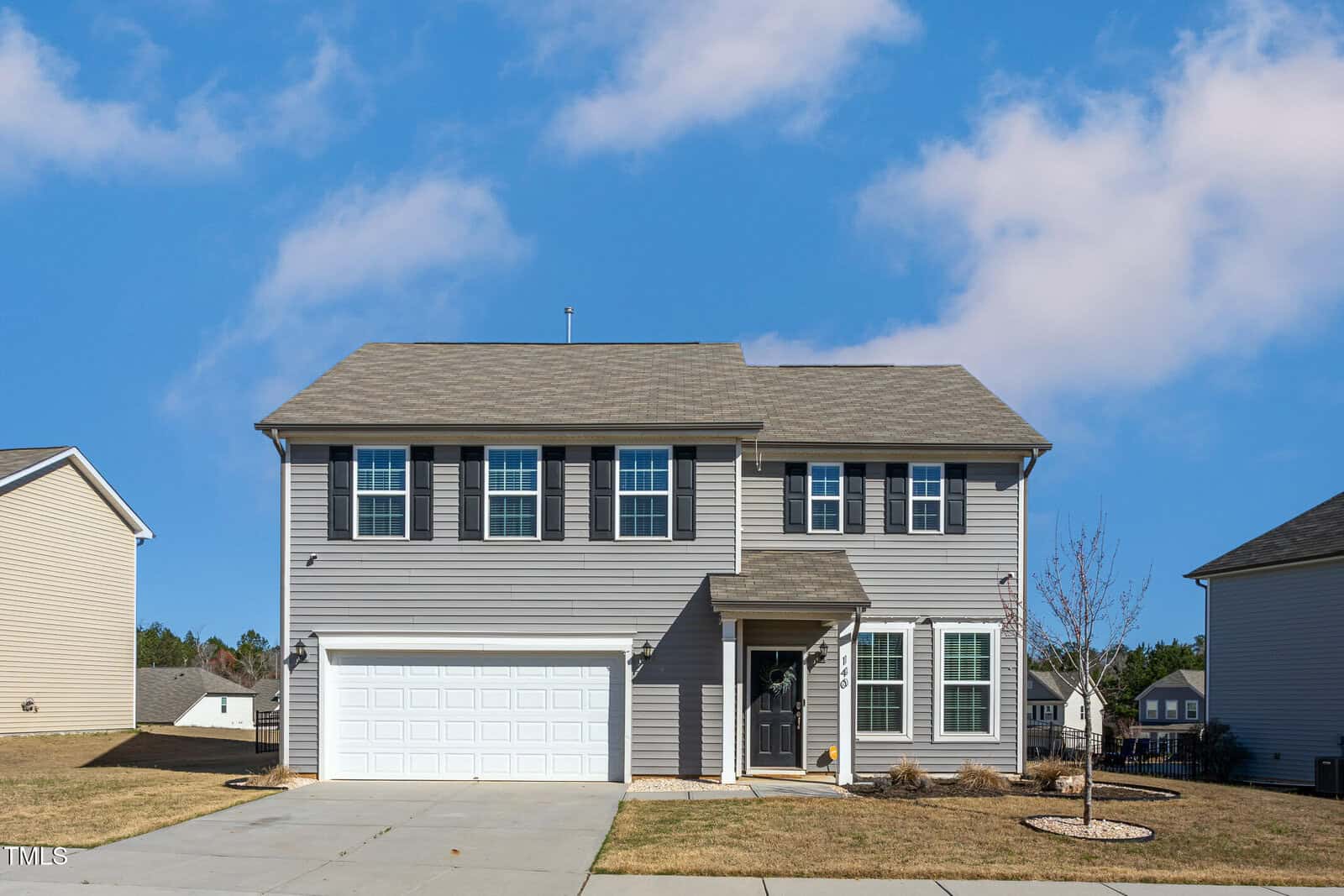 A two-story gray house with white trim and black shutters, featuring a double garage and a small front porch. The yard is mostly grass with a young tree and minimal landscaping under a blue sky with scattered clouds.