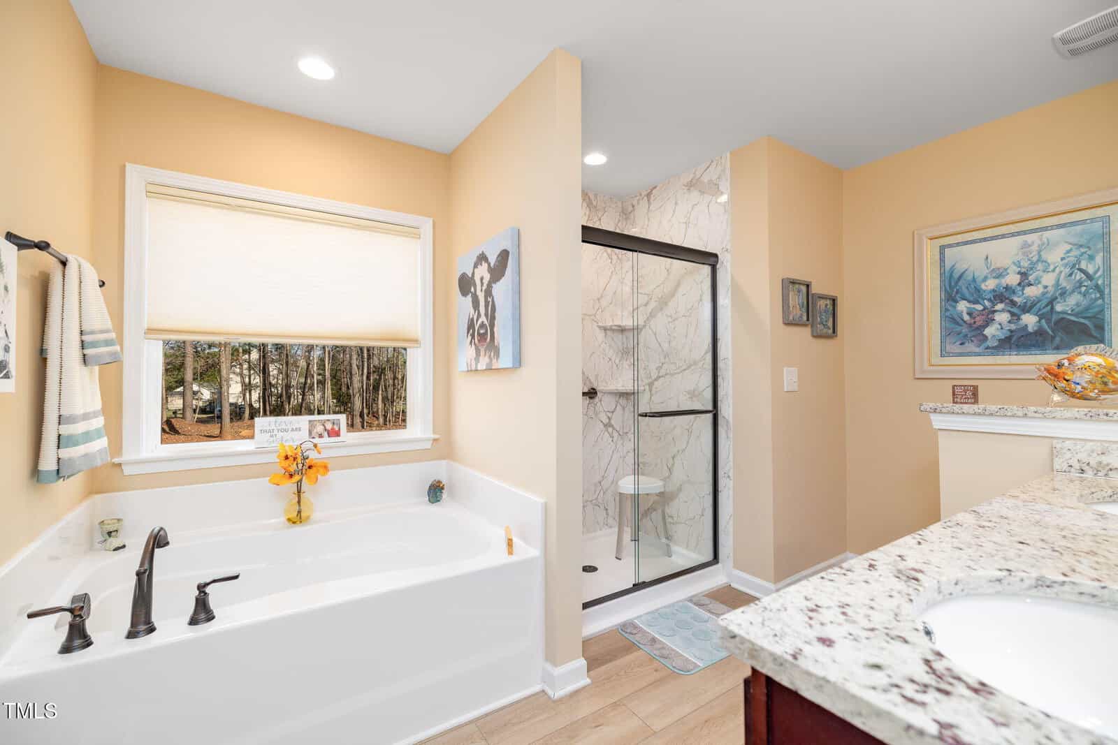 A bathroom with a white bathtub, black fixtures, a walk-in shower with glass door, granite countertop, and beige walls. There are framed pictures and a window with a view of trees. Towels hang on the wall.