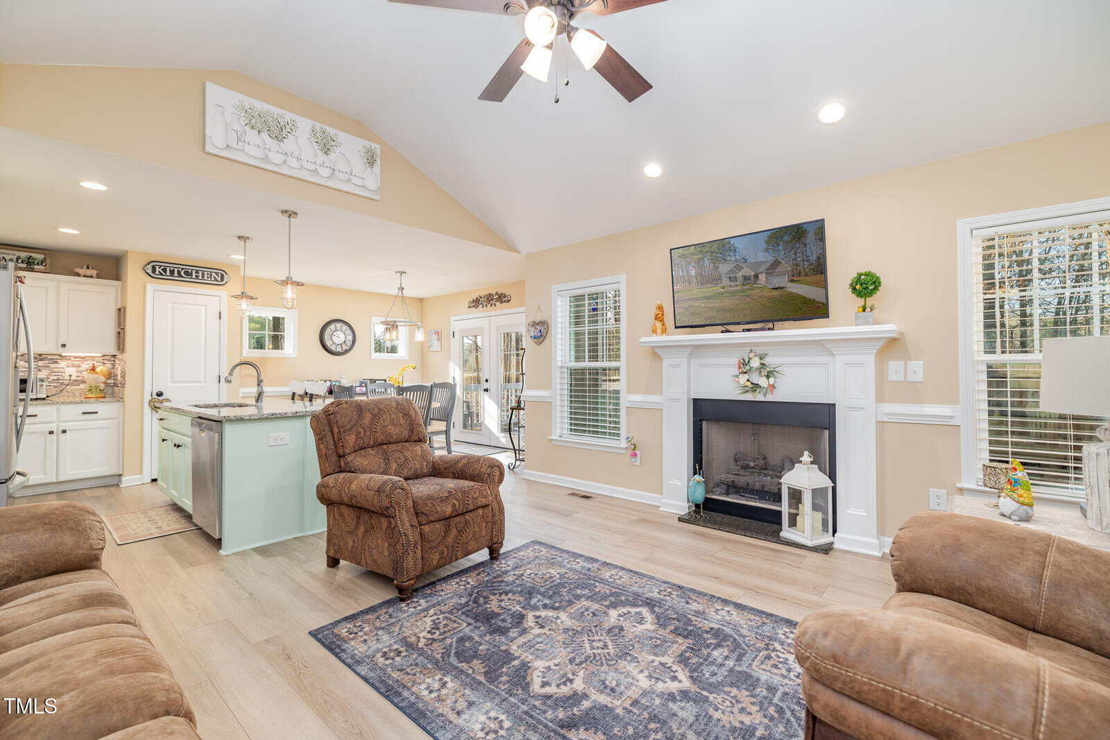 A bright living room with tan walls, wood floors, two brown armchairs, a fireplace with decor, a patterned rug, and an open view of the kitchen and dining area in the background. Ceiling fan and recessed lights above.