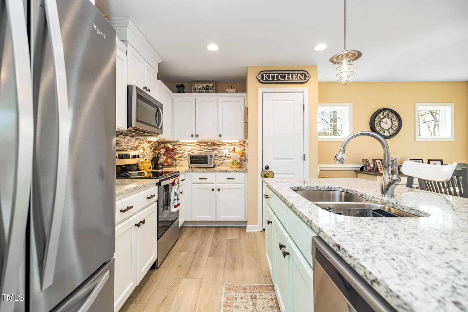 A modern kitchen with white cabinets, stainless steel appliances, granite countertops, a light wood floor, and a yellow accent wall. An island with a sink is in the foreground; decorative items and a "KITCHEN" sign are visible.