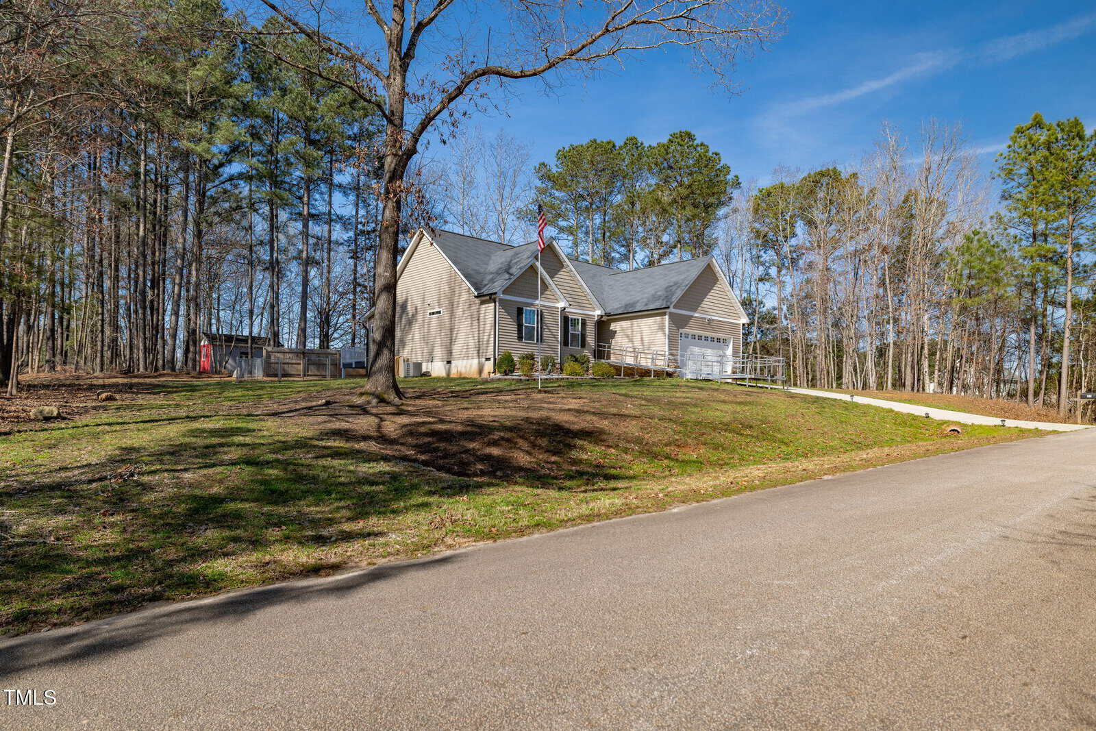 A single-story beige house with a gray roof sits on a spacious lot next to a paved road, surrounded by tall trees and a partially grassy yard under a clear blue sky.