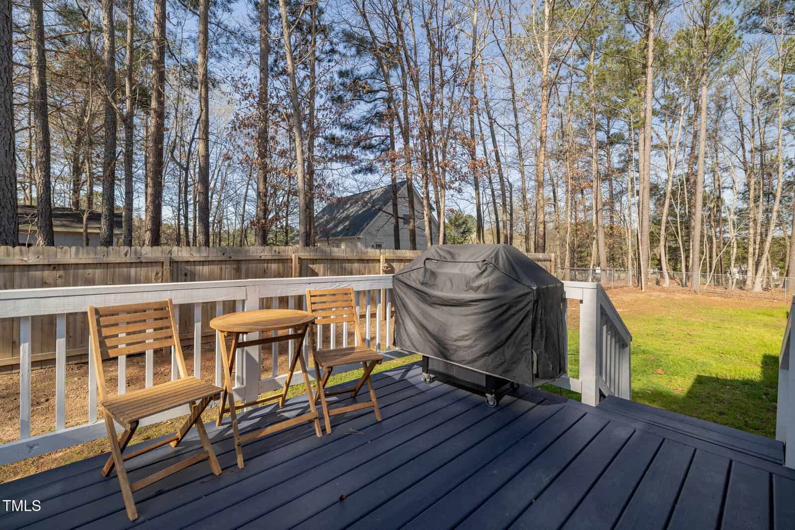 A small wooden deck with two chairs, a round table, and a covered grill. The deck overlooks a fenced backyard with trees and a clear sky.