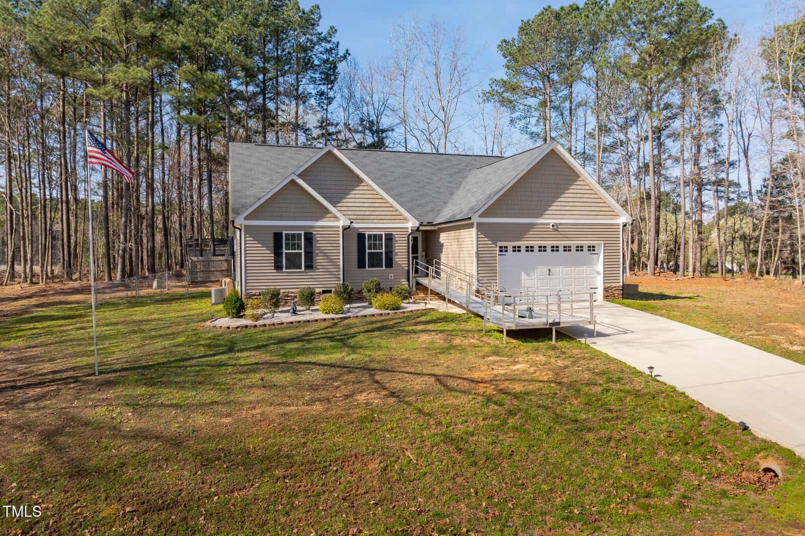 Single-story house with beige siding, gray roof, a covered front porch, and an attached two-car garage. A concrete driveway and ramp lead to the entrance. An American flag is on a flagpole in the grassy front yard, surrounded by tall trees.