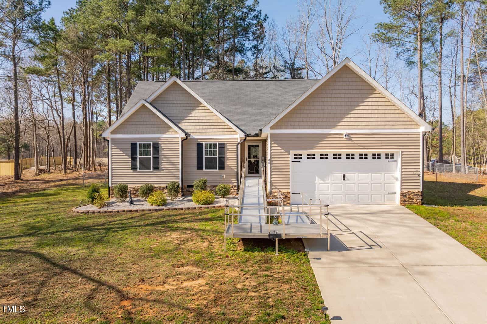 Single-story beige house with white trim and a gray shingled roof, featuring a two-car garage. A wheelchair-accessible ramp leads to the front door. The yard has minimal landscaping and trees in the background.