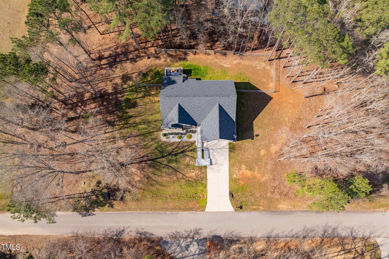 Aerial view of a single-family house with a gray roof, driveway, and front yard, surrounded by trees and bordered by a paved road. The property is located in a wooded area with sparse grass and fallen leaves.
