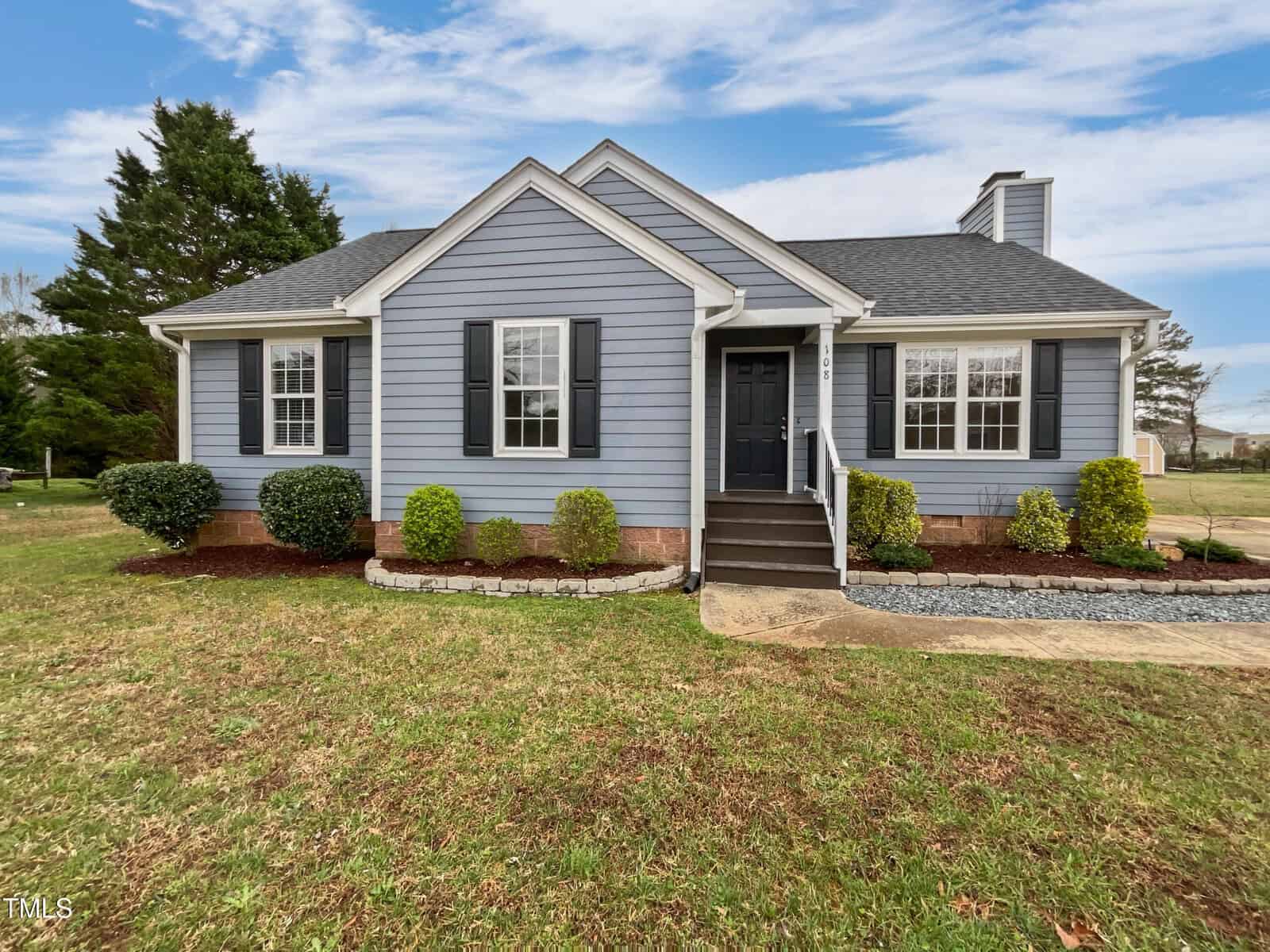 A single-story house with light blue siding, dark shutters, and a front porch with steps. Shrubs and mulch border the front, and the lawn is trimmed with trees and a partly cloudy sky in the background.