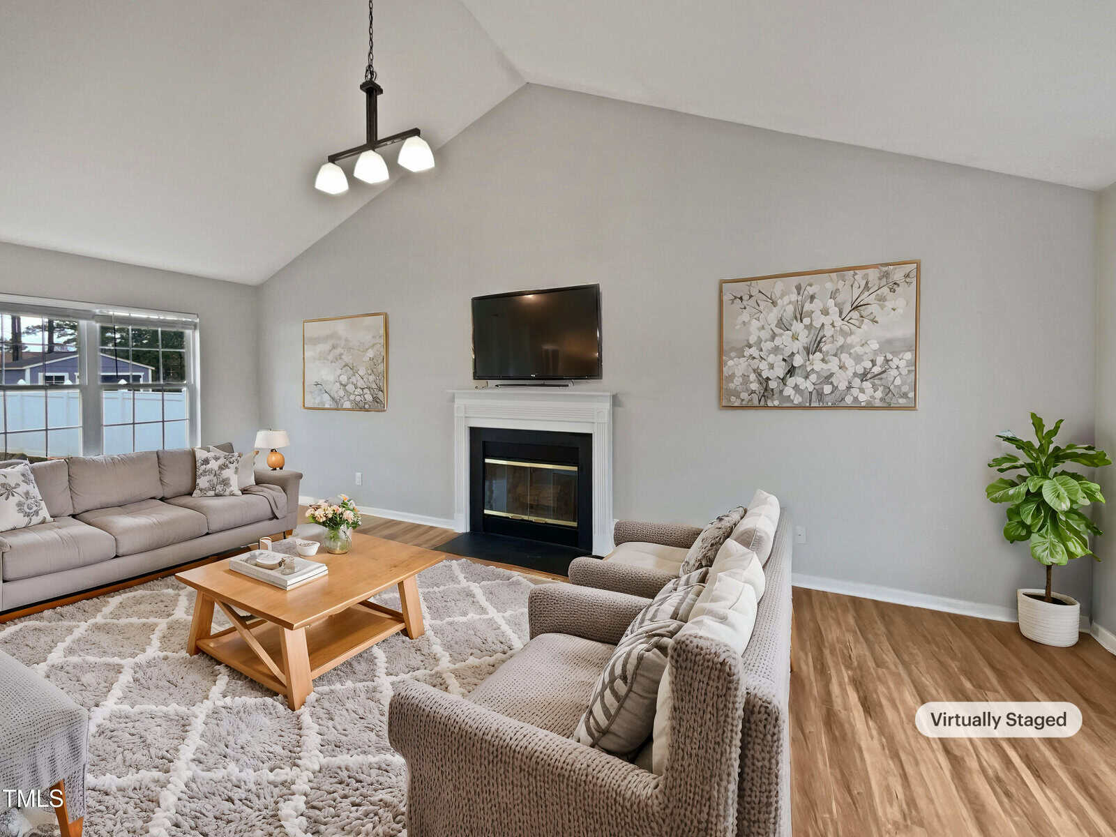 A living room with a vaulted ceiling, light gray walls, a fireplace with a TV above it, two beige armchairs, a beige sofa, a wooden coffee table, a shaggy rug, framed floral art, and a potted plant. "Virtually Staged" label in corner.