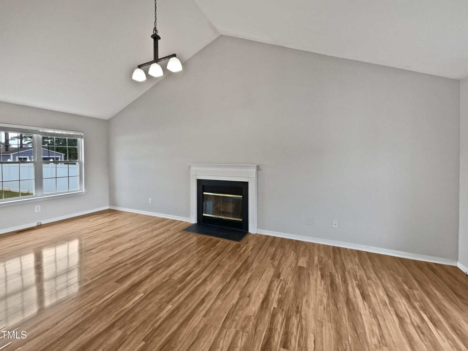 A spacious, empty living room with light wood flooring, light gray walls, a white fireplace with a black hearth, a large window, vaulted ceiling, and a black three-light chandelier hanging from the ceiling.