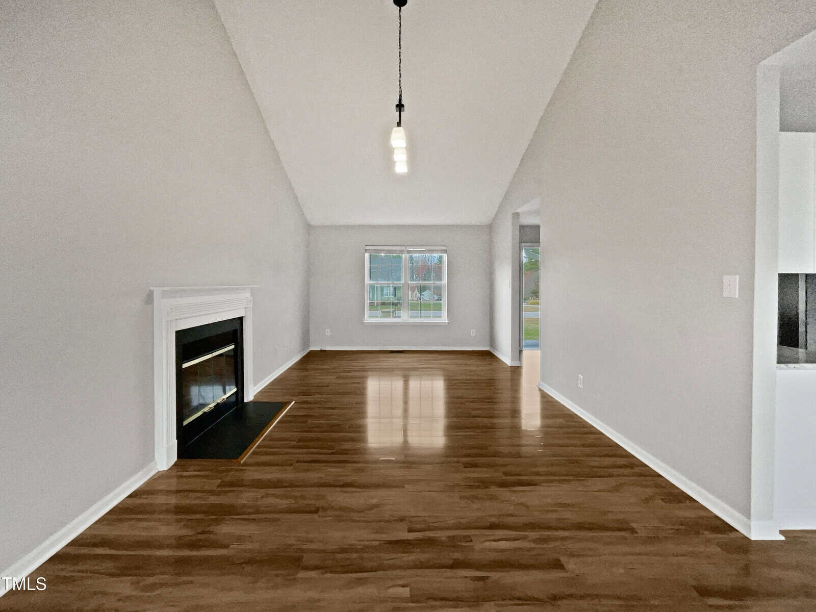 A spacious, empty living room with light gray walls, wood flooring, a white fireplace on the left, a large window at the back, pendant lights on the ceiling, and an open doorway leading to another room on the right.