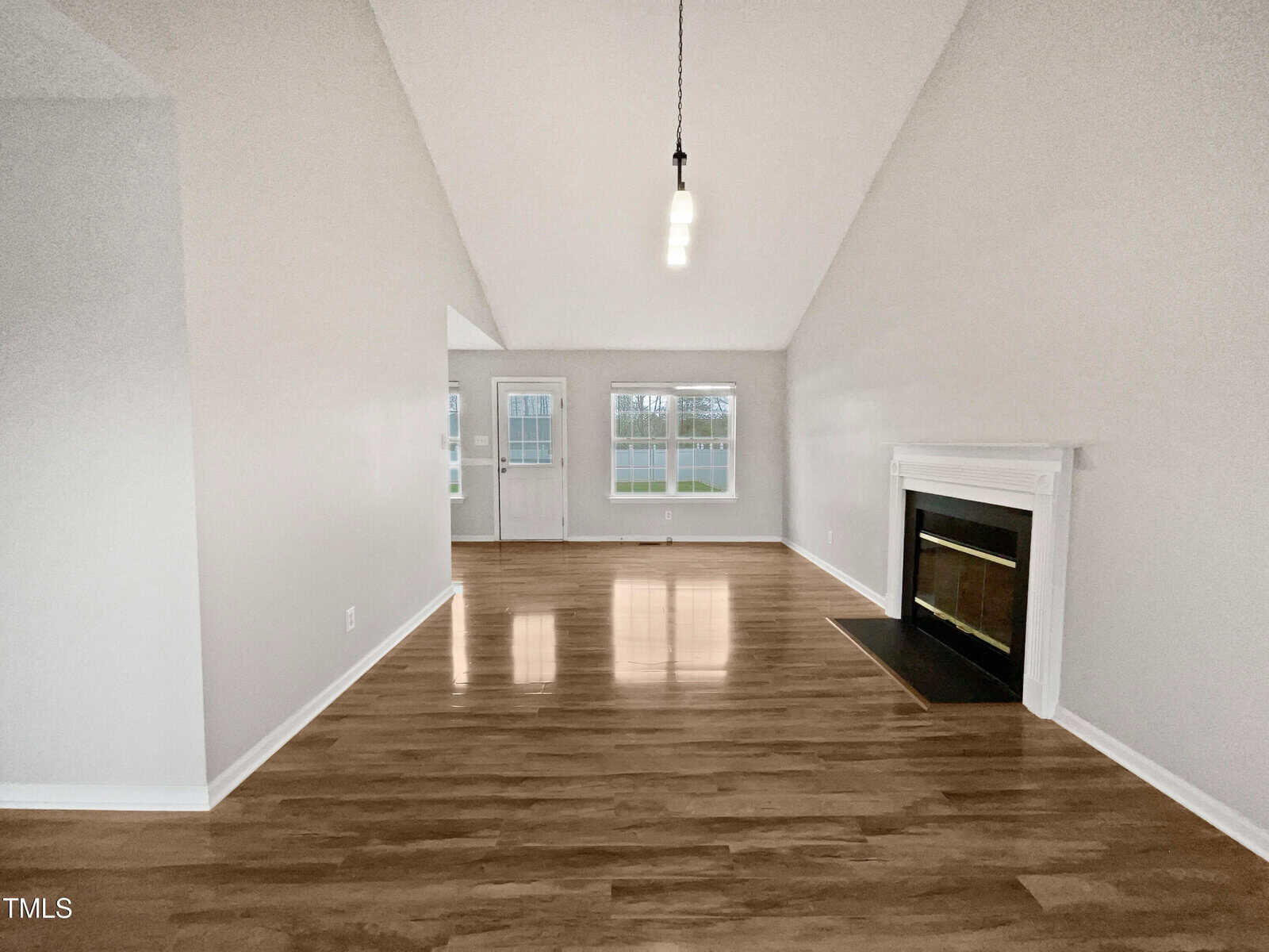 A spacious, empty living room with light gray walls, wood flooring, a white fireplace on the right, two hanging light fixtures, and large windows letting in natural light at the far end.
