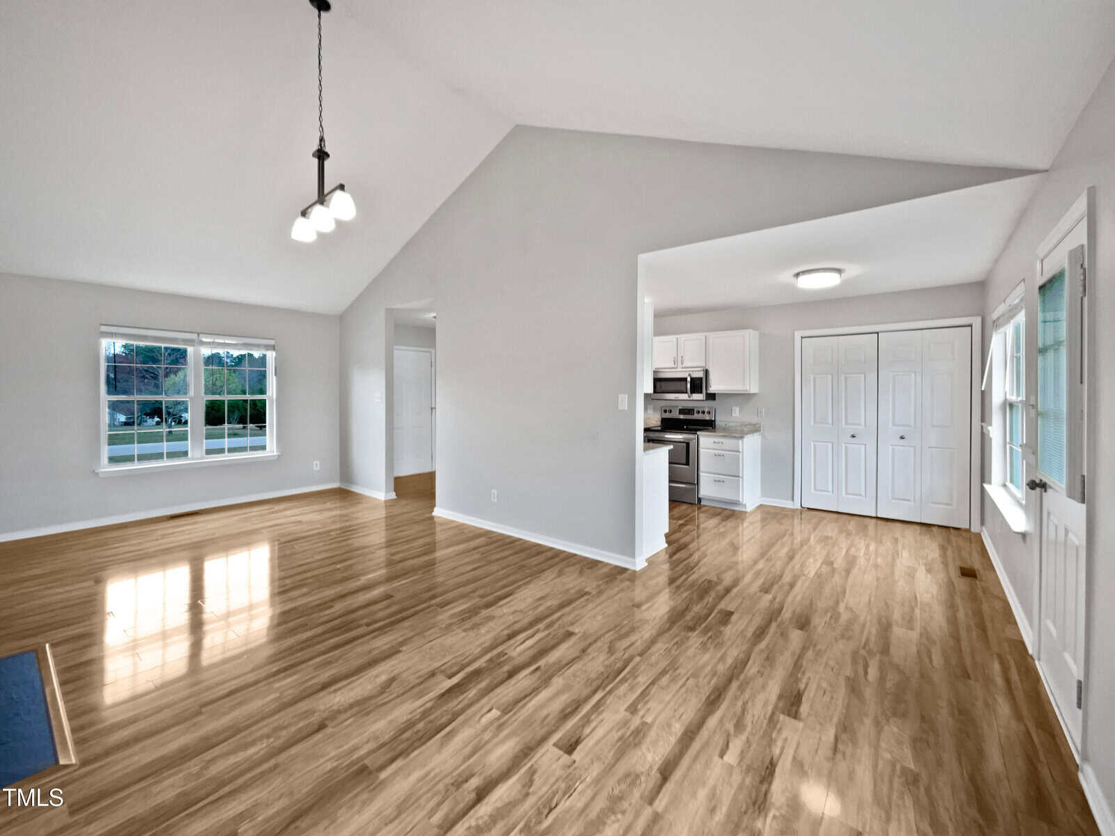 Open living area with light wood floors, white walls, and vaulted ceiling. Kitchen with white cabinets and stainless appliances visible through open doorway. Large windows let in natural light.