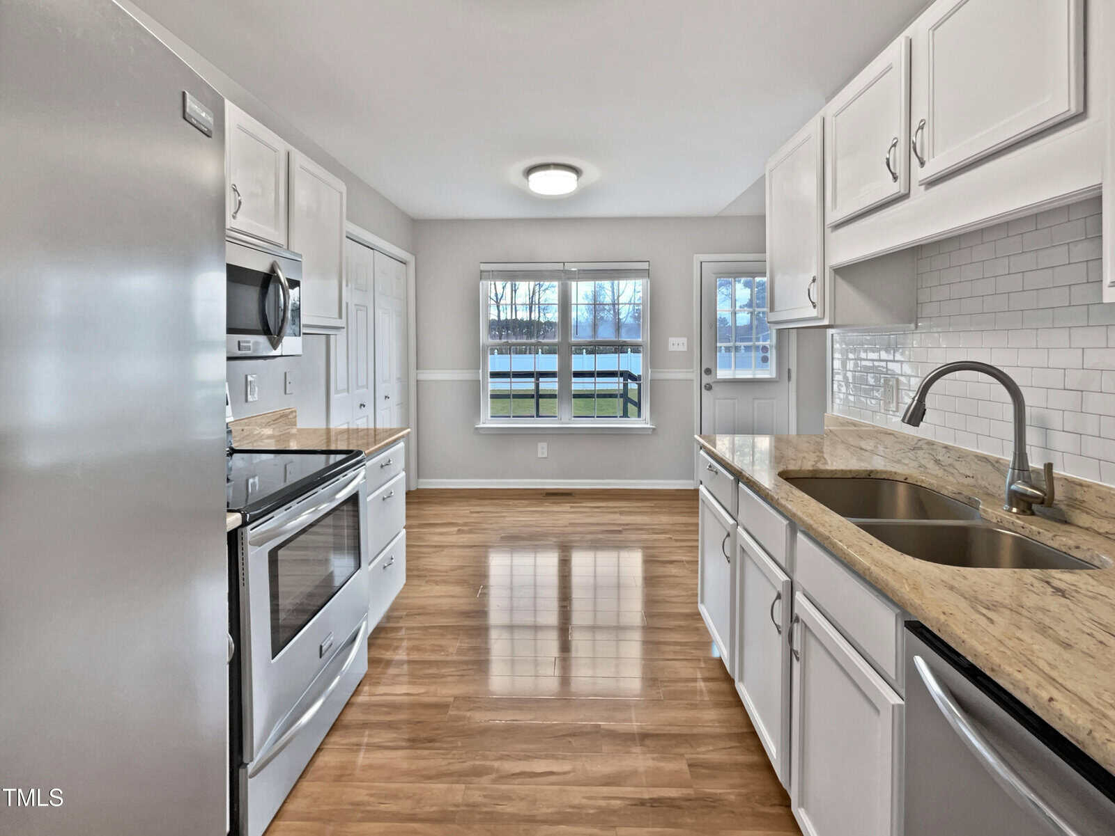 Modern kitchen with white cabinets, stainless steel appliances, a double sink, and light granite countertops. The floor is wood, and large windows in the adjacent room let in natural light. Walls are light gray with white trim.