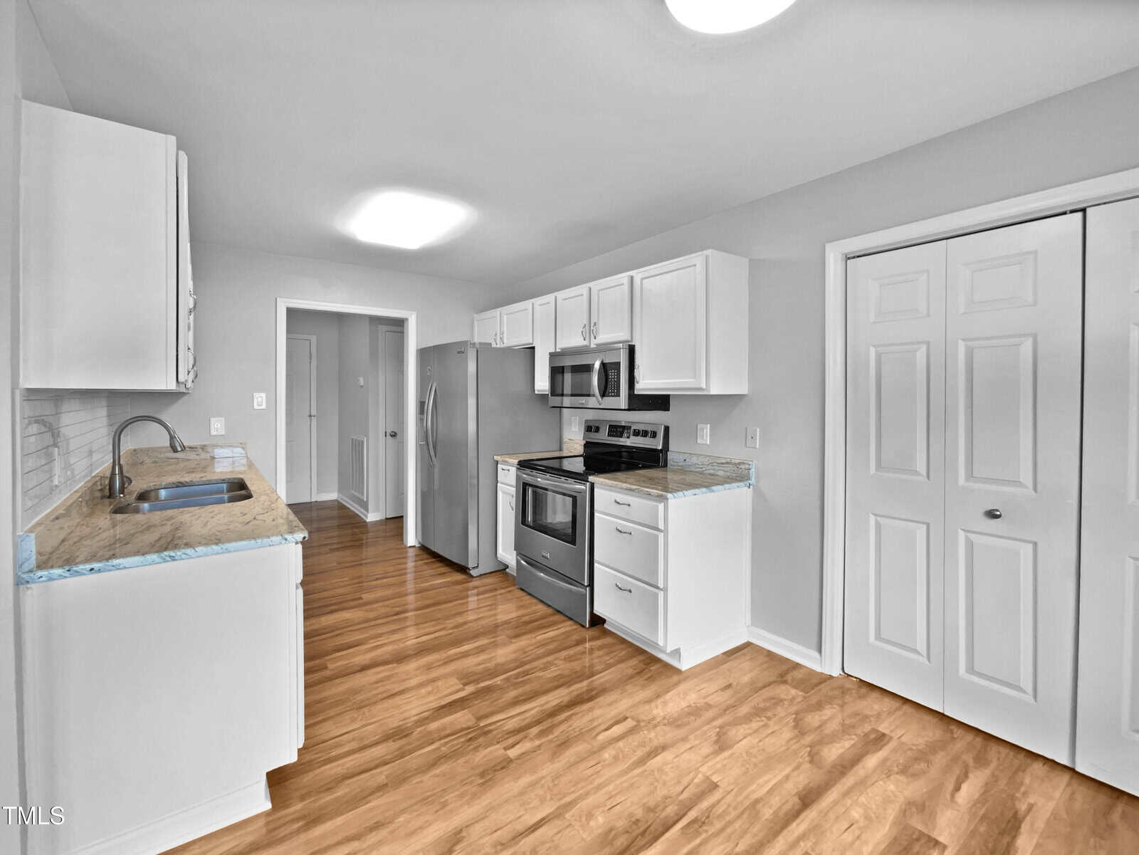 Modern kitchen with white cabinets, stainless steel appliances, marble countertops, wood flooring, under-cabinet sink, and double pantry doors. Room is well-lit with overhead lights. Walls and ceiling are painted white.
