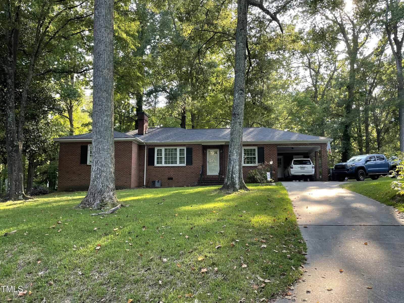A single-story brick house at 321 Willowood Drive boasts a charming front porch amidst towering trees. Two vehicles rest in the driveway to the right, while a grassy lawn with scattered leaves complements its surroundings. The backdrop features a serene wooded area in Henderson.