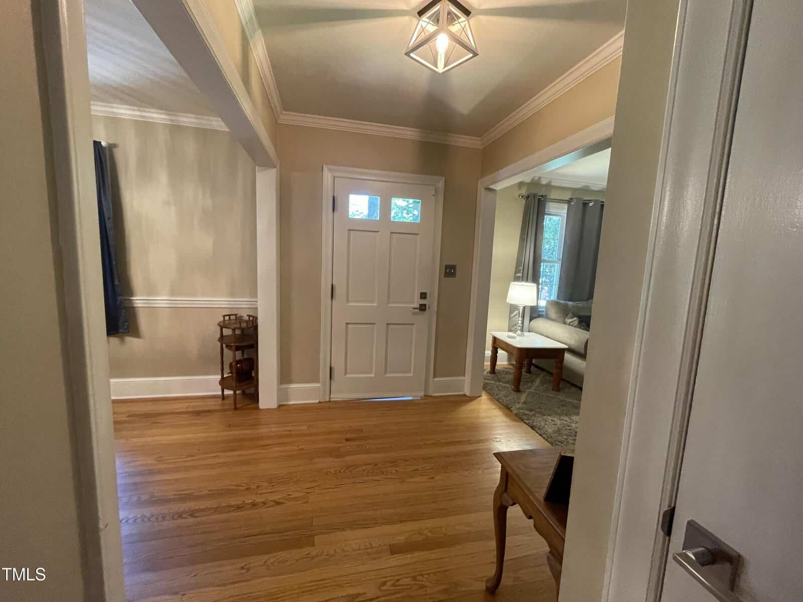A hallway with wooden flooring and beige walls leads to a front door with a window. A small table is on the right, and a room with a couch and lamp is visible through an archway on the right. A decorative light fixture hangs from the ceiling.