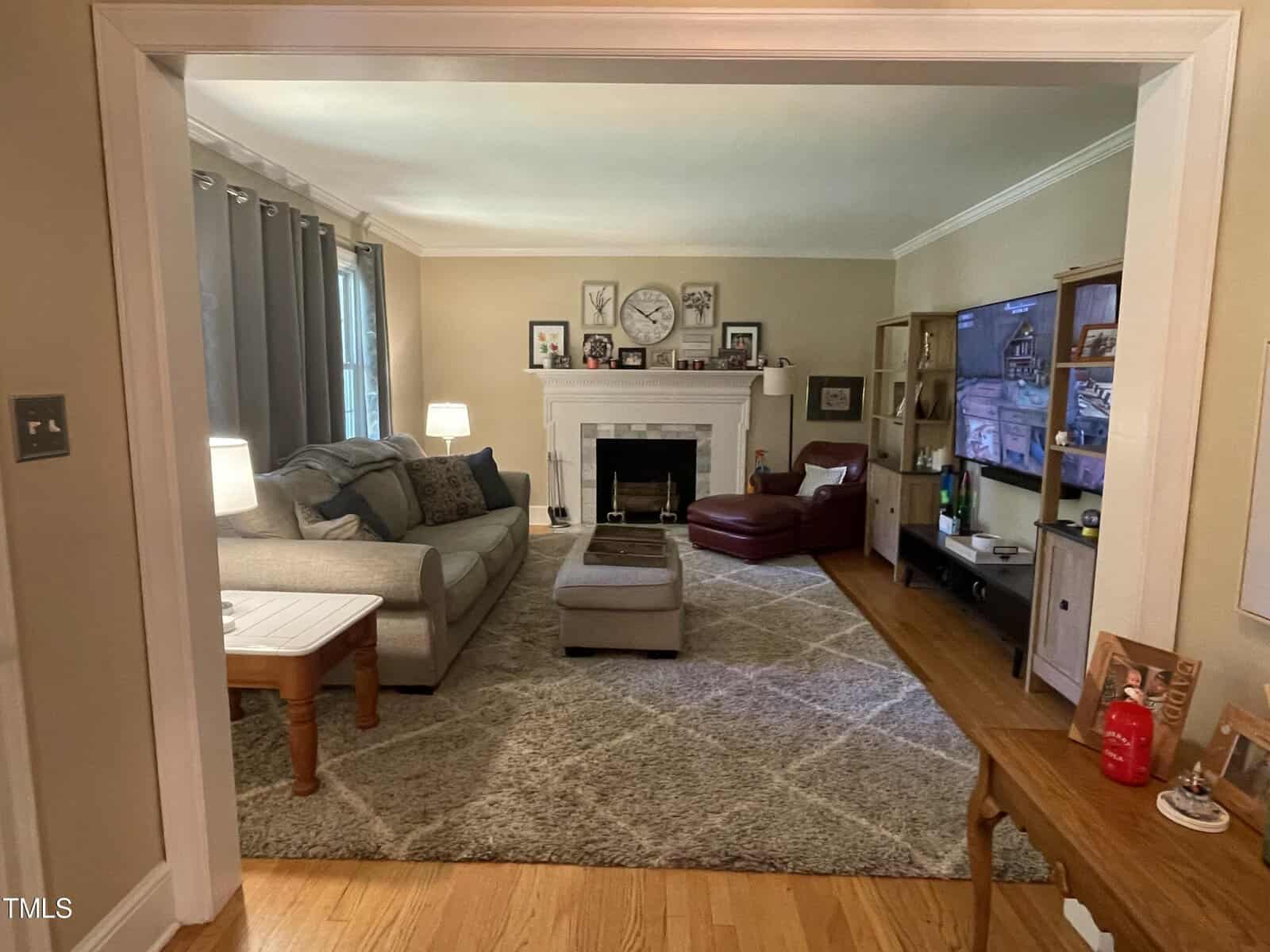A cozy living room with a gray sectional sofa, a red armchair, and a patterned rug. The fireplace mantel is decorated with a clock and framed photos. A large TV is mounted on a wooden cabinet. The room is well-lit with lamps and natural light.