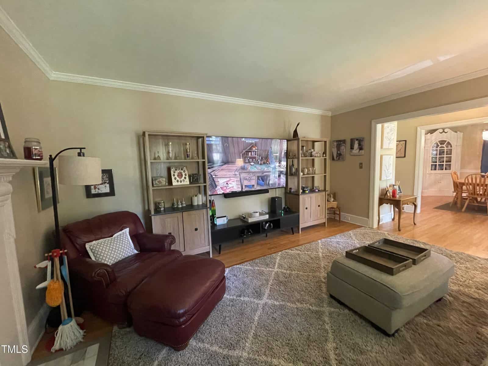 A living room with a large TV centered on a stand between two cabinets. A red leather armchair is next to a white lamp, and a cushioned ottoman sits on a gray rug. The room connects to a dining area with a wooden table and chairs.