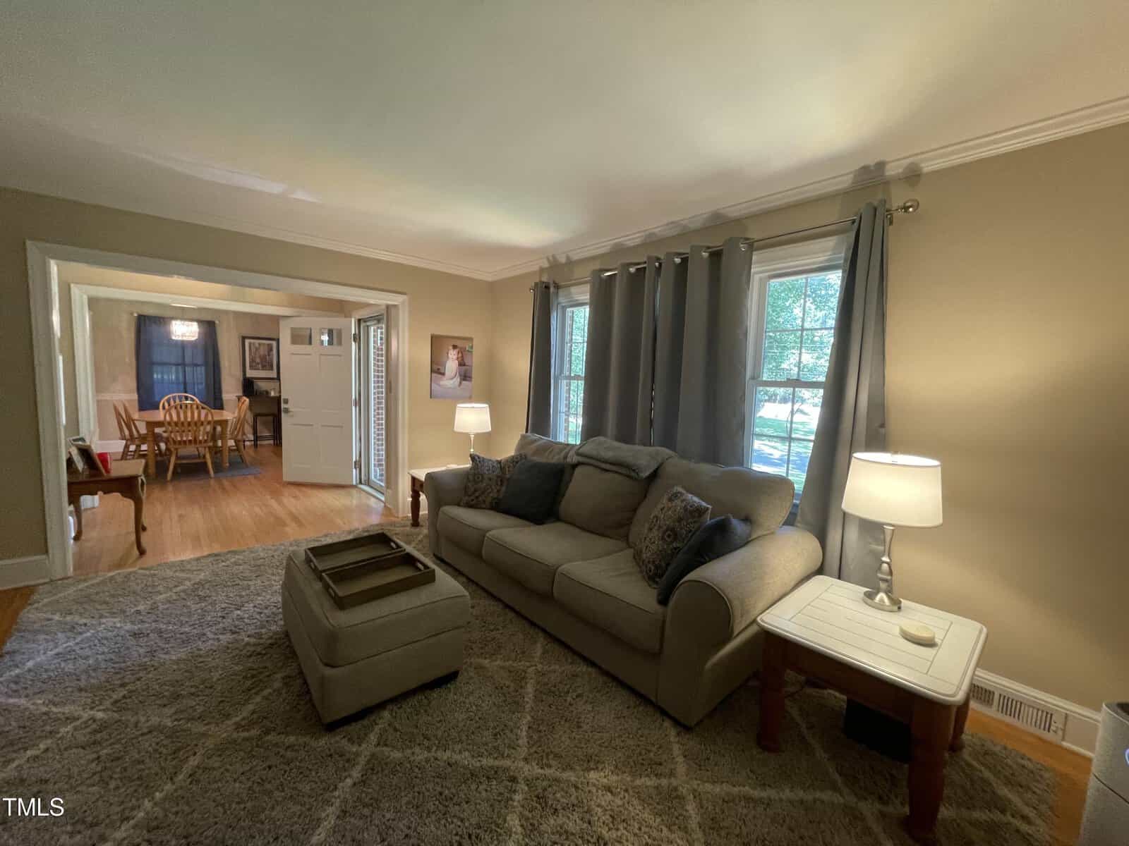 A living room with a grey sofa and matching ottoman on a patterned rug. Two lamps on side tables flank the sofa. Curtains frame a window. A doorway leads to a dining area with a wooden table and chairs. Walls are painted beige.