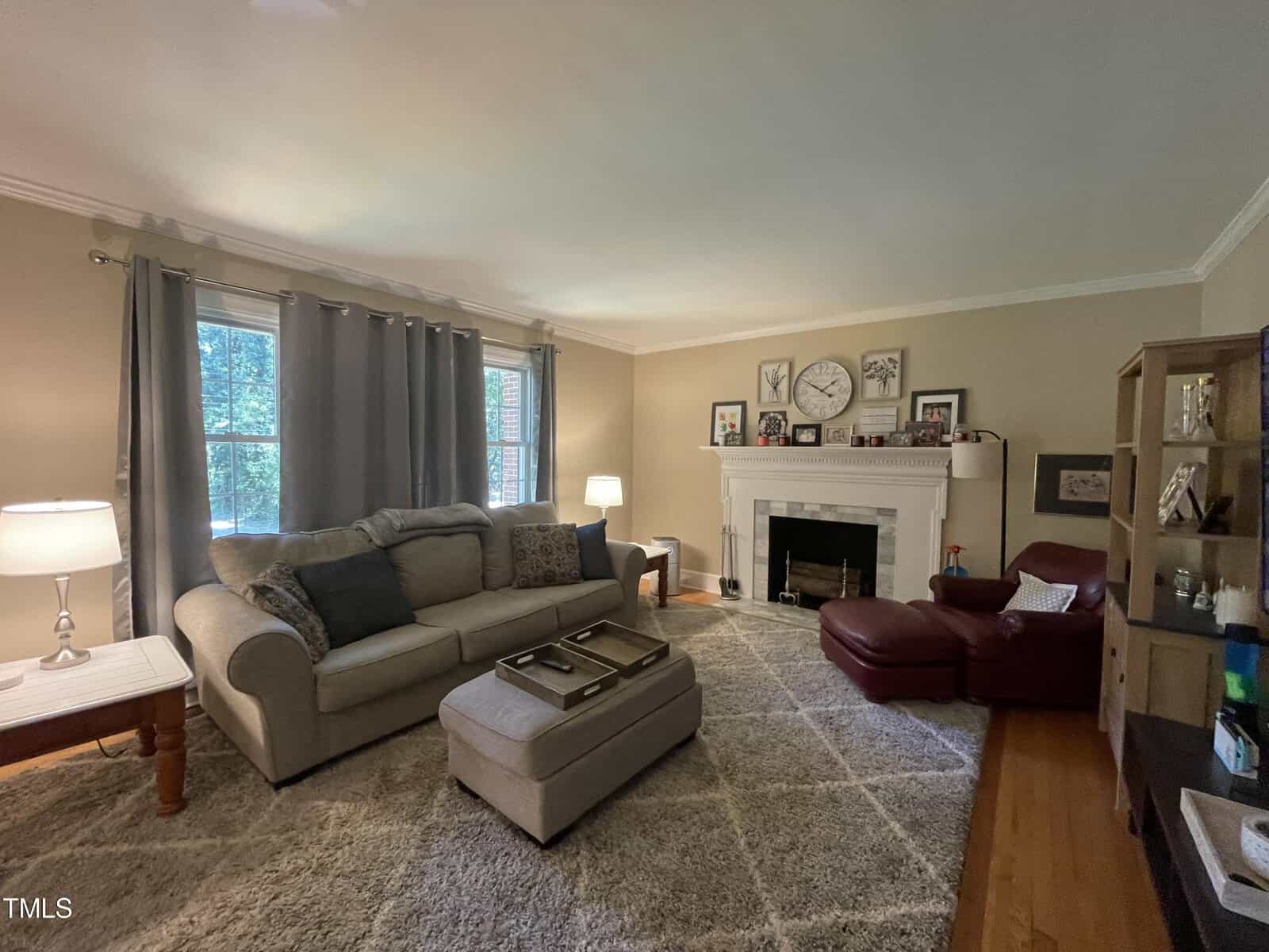 A cozy living room with a large gray sofa and a matching ottoman. A red leather chair and ottoman sit by a white fireplace. The walls feature framed pictures and a large clock. Two lamps and a bookshelf are also present. A soft rug covers the floor.