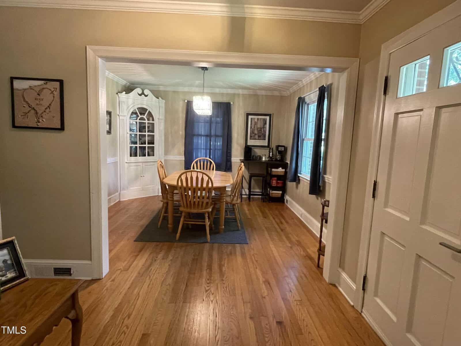 A dining room with a wooden table and six chairs on a rug. There's a cabinet in the corner, large windows with curtains, and walls adorned with framed pictures. A chandelier hangs over the table, and hardwood floors run throughout the room.
