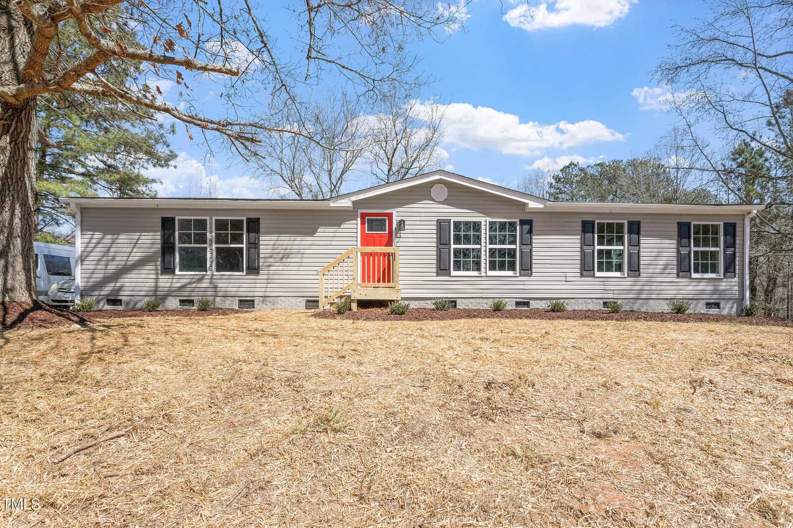 A single-story, light gray manufactured home with white trim, a bright red front door, small front porch with steps, and a large front yard with straw-covered ground and minimal landscaping. Trees and blue sky in the background.