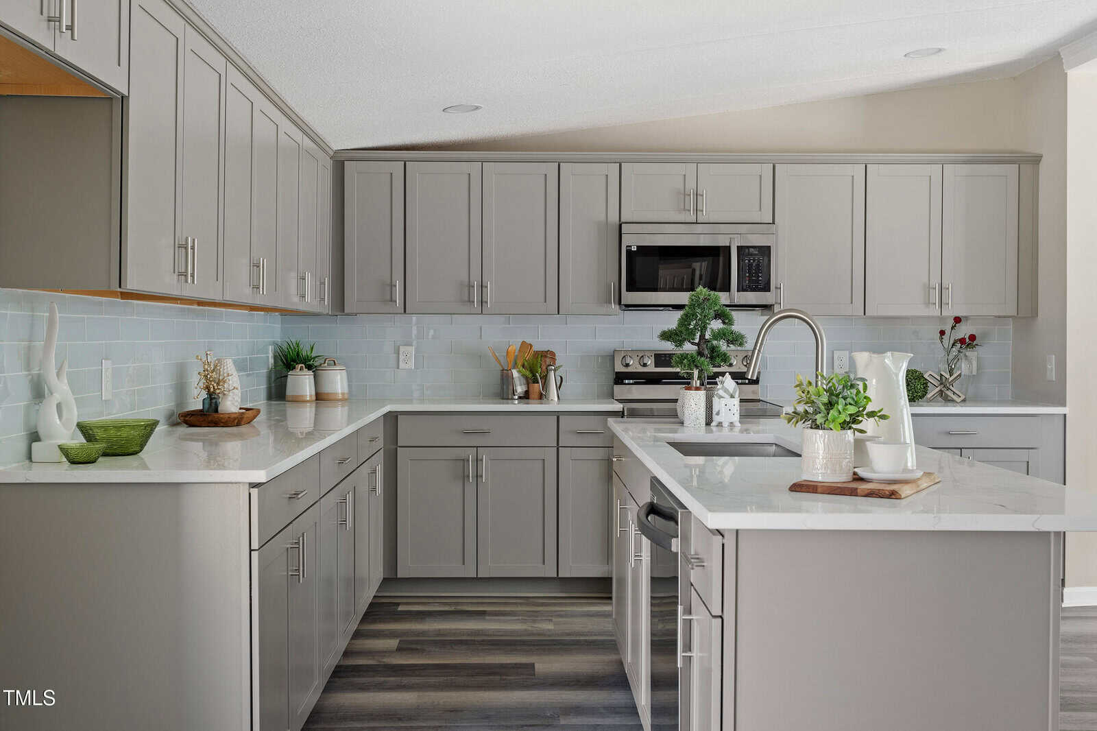 A modern kitchen with gray cabinets, white countertops, a stainless steel stove and microwave, a sink on the island, and decorative plants and kitchenware on the counters. Light blue tile backsplash and wood-look flooring.