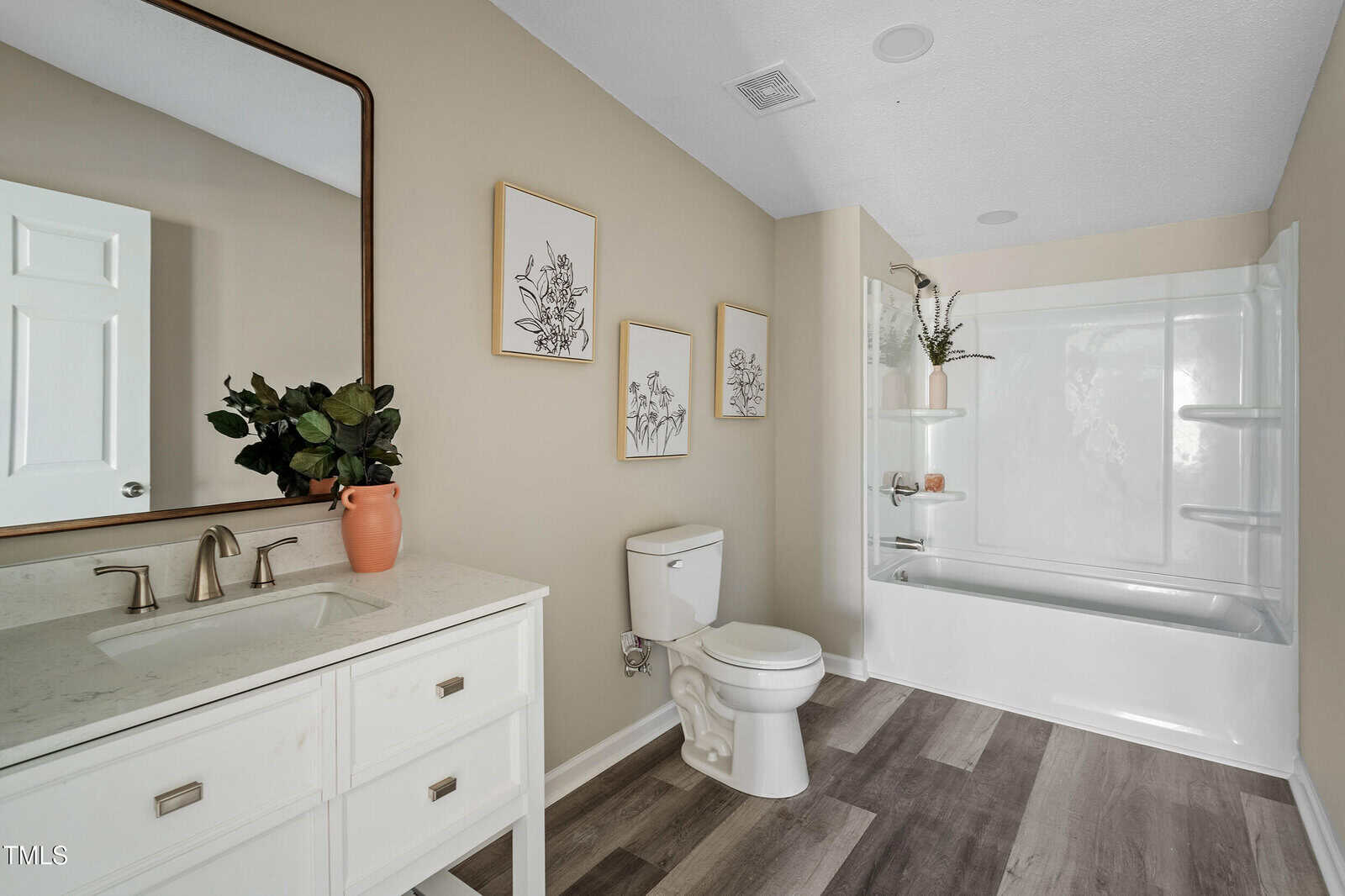 Modern bathroom with beige walls, white vanity and sink, wall mirror, toilet, three framed art pieces, potted plant, and a bathtub-shower combo with built-in shelves and a small plant on the ledge. Light wood flooring throughout.