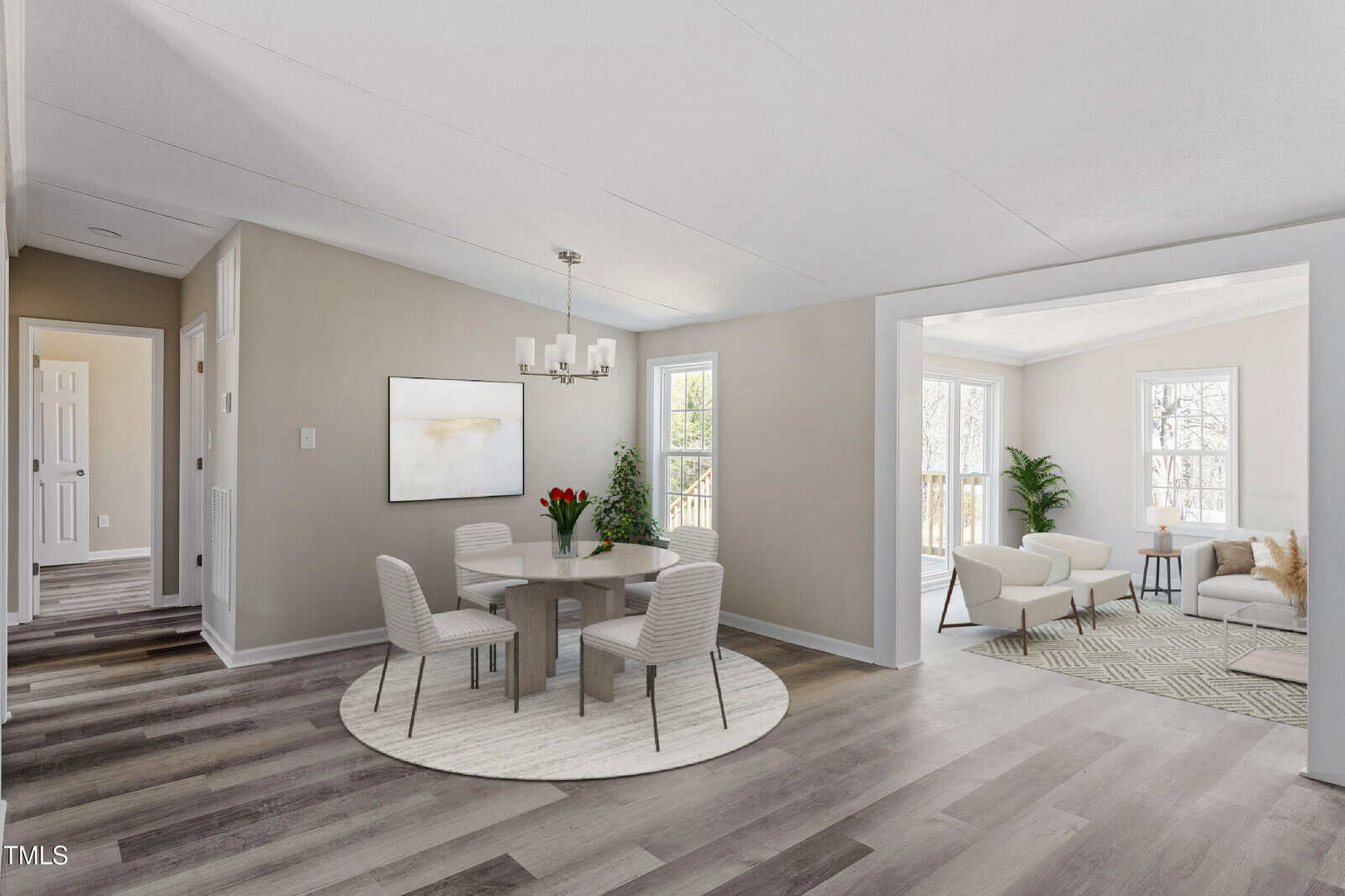 A dining room with a round table and four chairs on a rug, light wood flooring, neutral walls, and a chandelier. An open doorway leads to a bright living area with windows, white chairs, and a green plant.