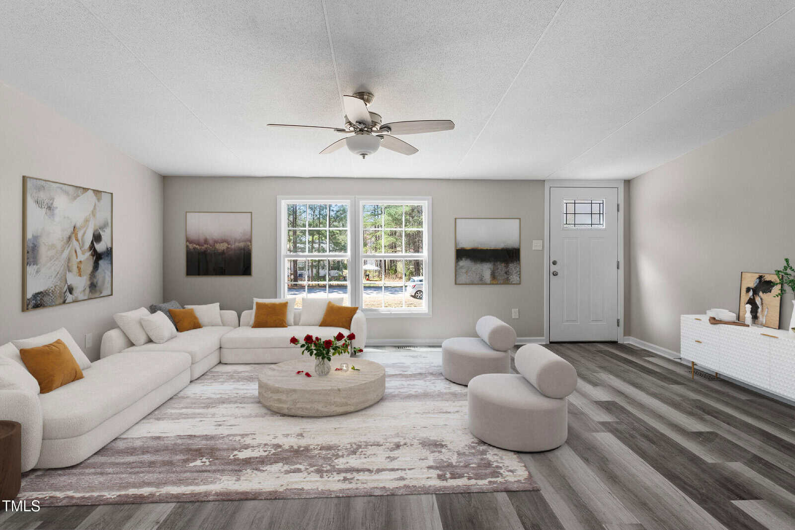 A modern living room with a white sectional sofa, circular chairs, a round coffee table with flowers, abstract wall art, a rug, wood-look flooring, a ceiling fan, and a door by a window letting in natural light.