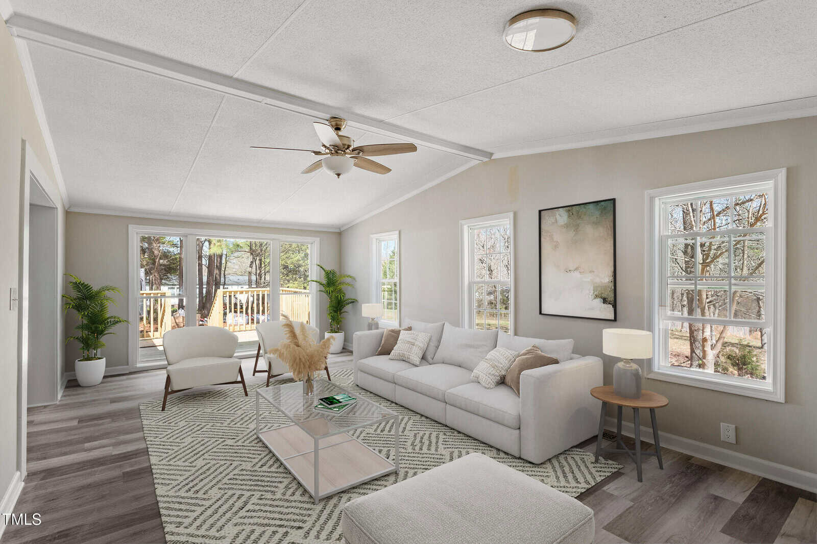 A bright living room with large windows, neutral walls, and wood flooring. The room is furnished with a white sofa, a glass coffee table, a patterned rug, two side tables, and a ceiling fan. A deck is visible outside.