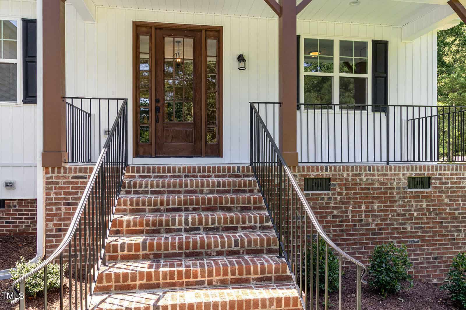 A brick staircase with black metal railings leads to a wooden front door with glass panels on a white house with vertical siding. There is a black outdoor light beside the door and a window on the right.