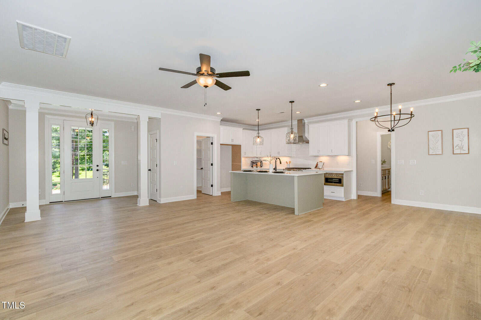 Open-concept living area with light wood flooring, white walls, ceiling fans, a kitchen with white cabinets and an island, two chandeliers, and double glass doors leading outside.