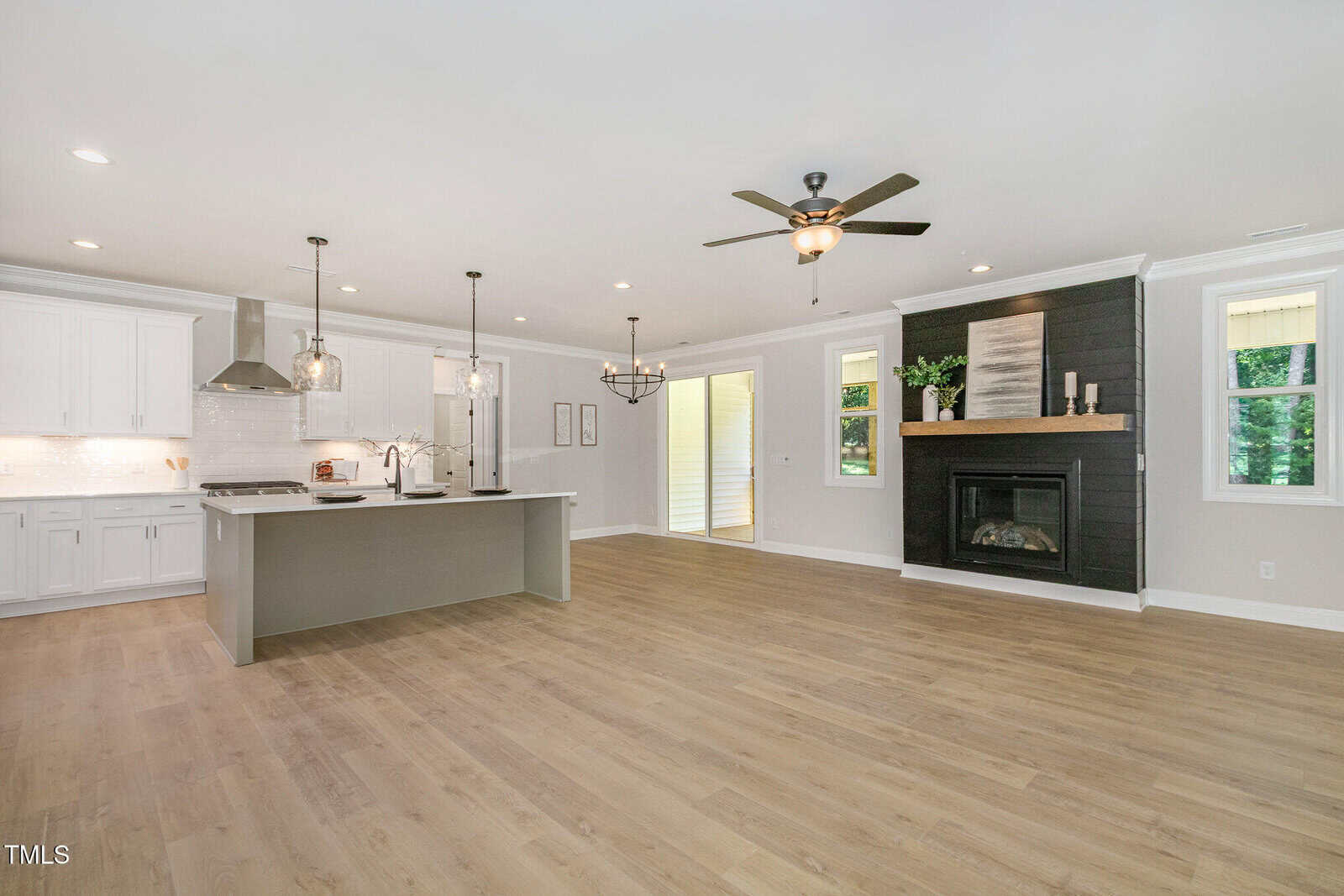 A spacious open-concept kitchen and living area with light wood flooring, white cabinets, a large island, stainless steel appliances, and a black fireplace with wood mantle. Ceiling fan and large windows provide natural light.