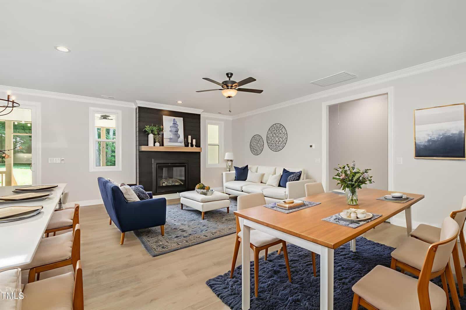 A modern open-concept living and dining area with a fireplace, blue and white furniture, wooden dining table with chairs, and decorative accents on the walls and table. Light wood flooring and natural light from windows.