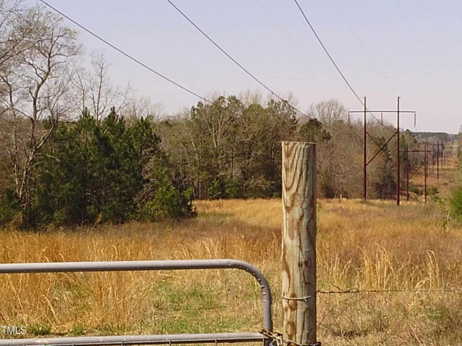 A rural landscape featuring a wooden fence post and metal gate in the foreground. In the background, there are tall power lines running through a grassy field with sparse trees under a clear sky.