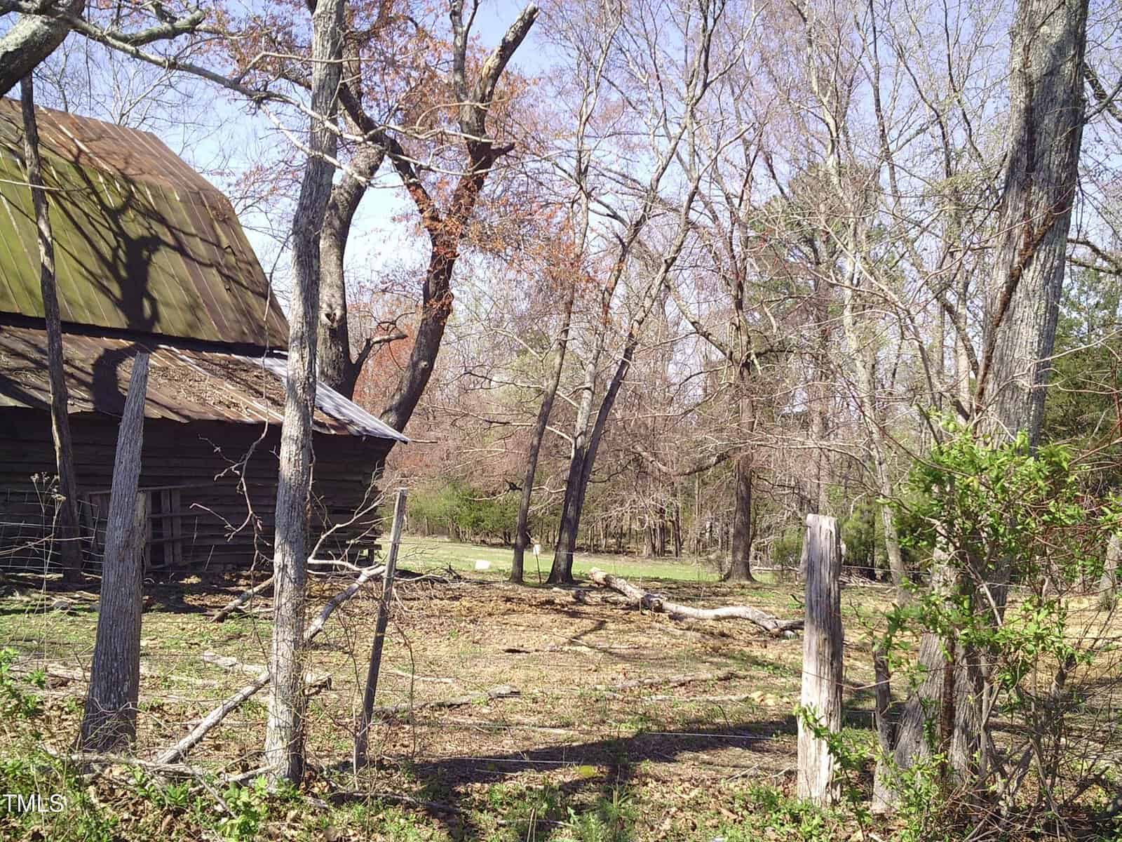Rural scene with a weathered, brown barn on the left, surrounded by bare trees. A rustic wooden fence runs along the foreground, with fallen branches scattered on the ground. The sky is clear and the foliage is sparse.