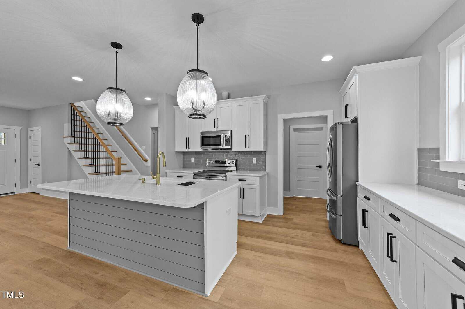 A modern kitchen with white cabinets, stainless steel appliances, a large island with a sink and gold faucet, two pendant lights, light wood flooring, and gray subway tile backsplash.