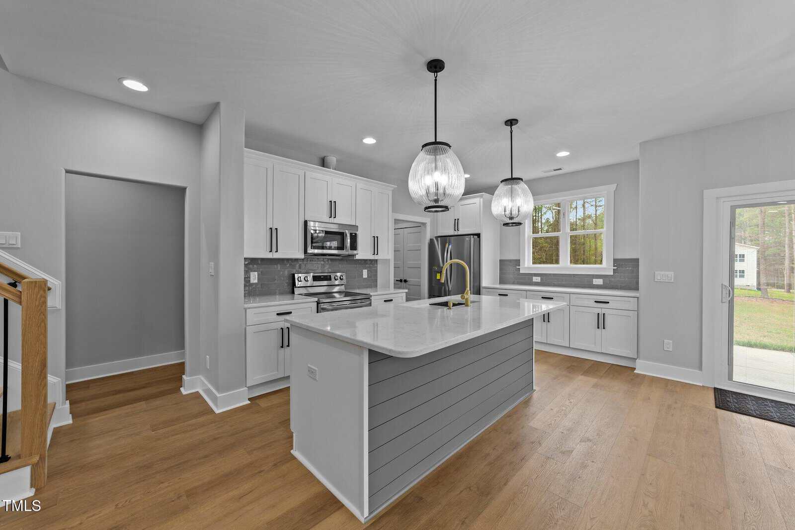 A modern kitchen with white cabinets, a gray island with a white countertop, stainless steel appliances, two pendant lights, wood flooring, and a view of a backyard through a glass door.