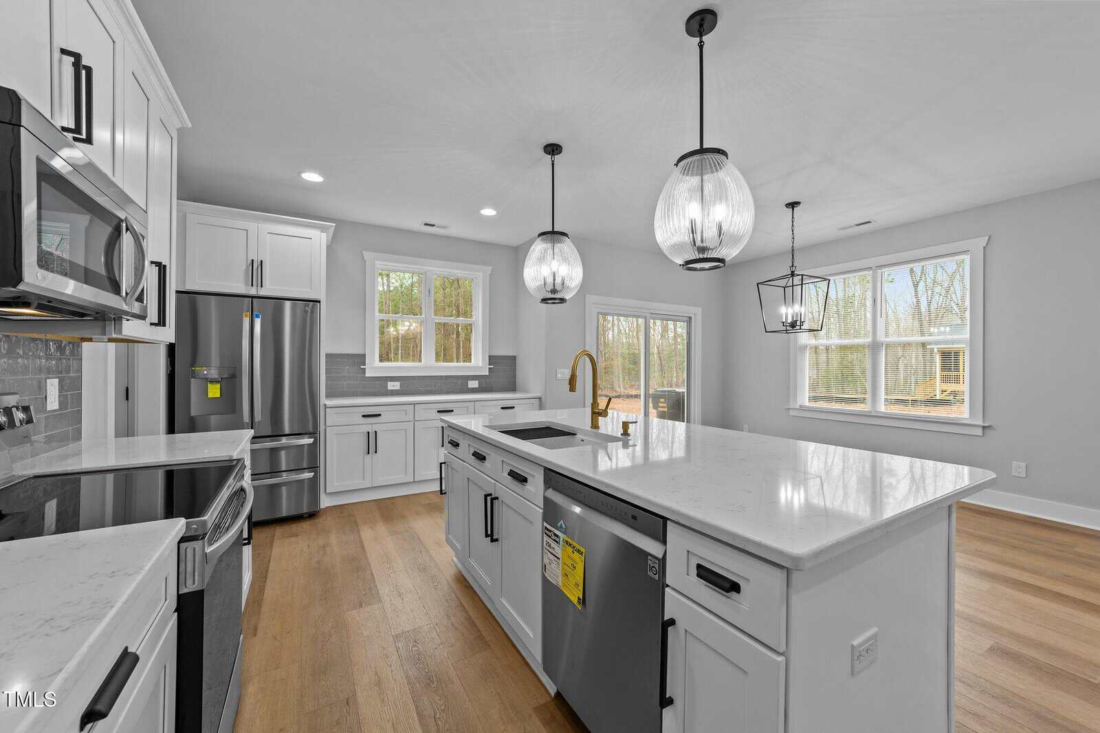 Modern kitchen with white cabinets, stainless steel appliances, a large island with built-in sink, pendant lighting, light wood floors, and multiple large windows allowing natural light into the space.