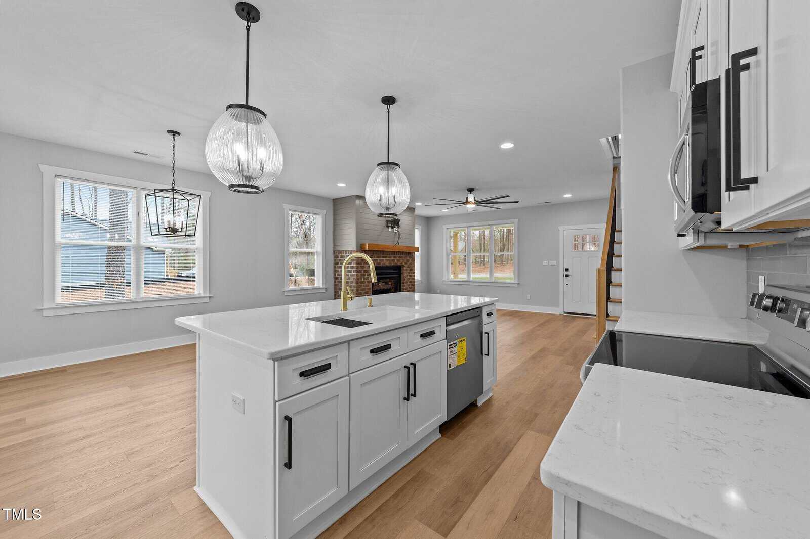Modern kitchen with white cabinets, marble countertop island, pendant lights, stainless steel appliances, and open layout extending to a living area with fireplace, large windows, ceiling fan, and wood flooring throughout.
