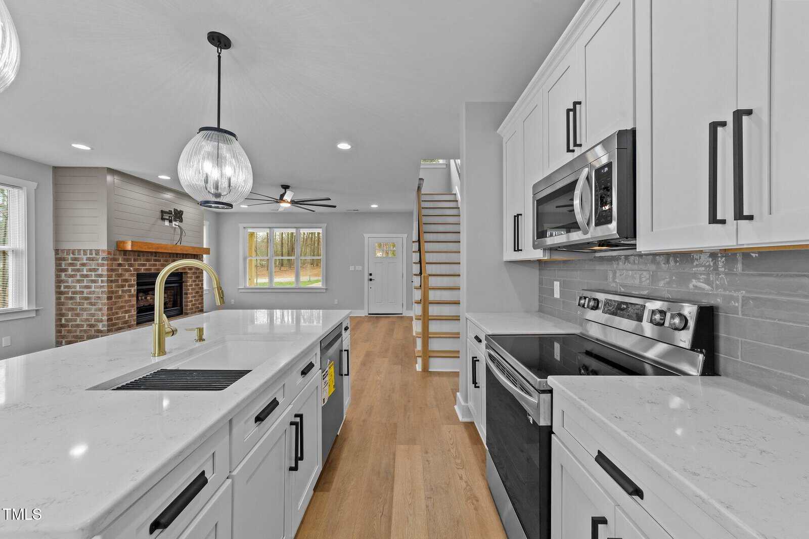 Modern kitchen with white cabinets, stainless steel appliances, light wood flooring, a large island with a sink, pendant lighting, and a view of a living area with a brick fireplace and staircase leading upstairs.