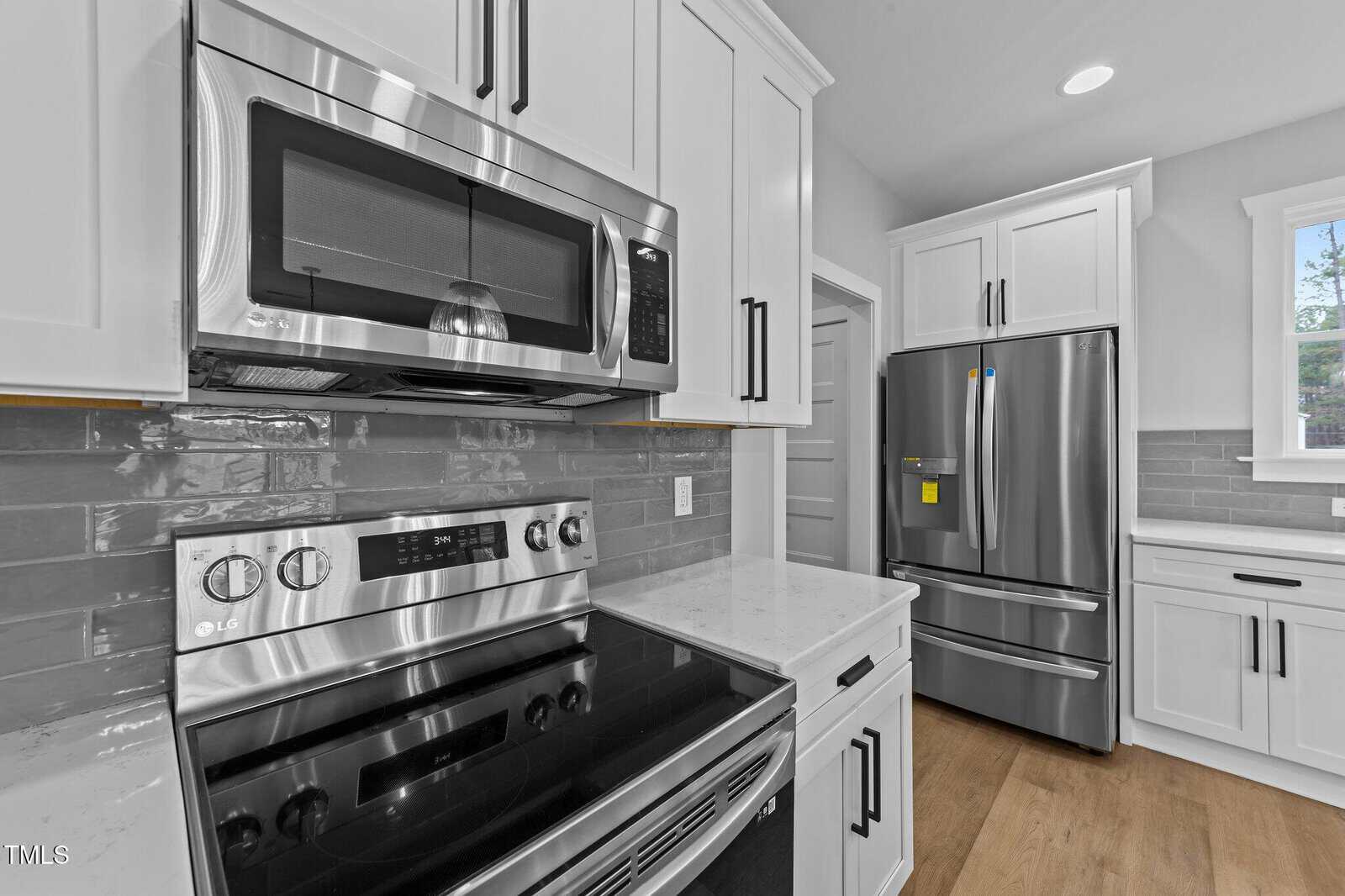 Modern kitchen with white cabinets, stainless steel microwave, oven, and refrigerator. Gray subway tile backsplash and wood floors are visible. A window lets in natural light above the counter on the right.