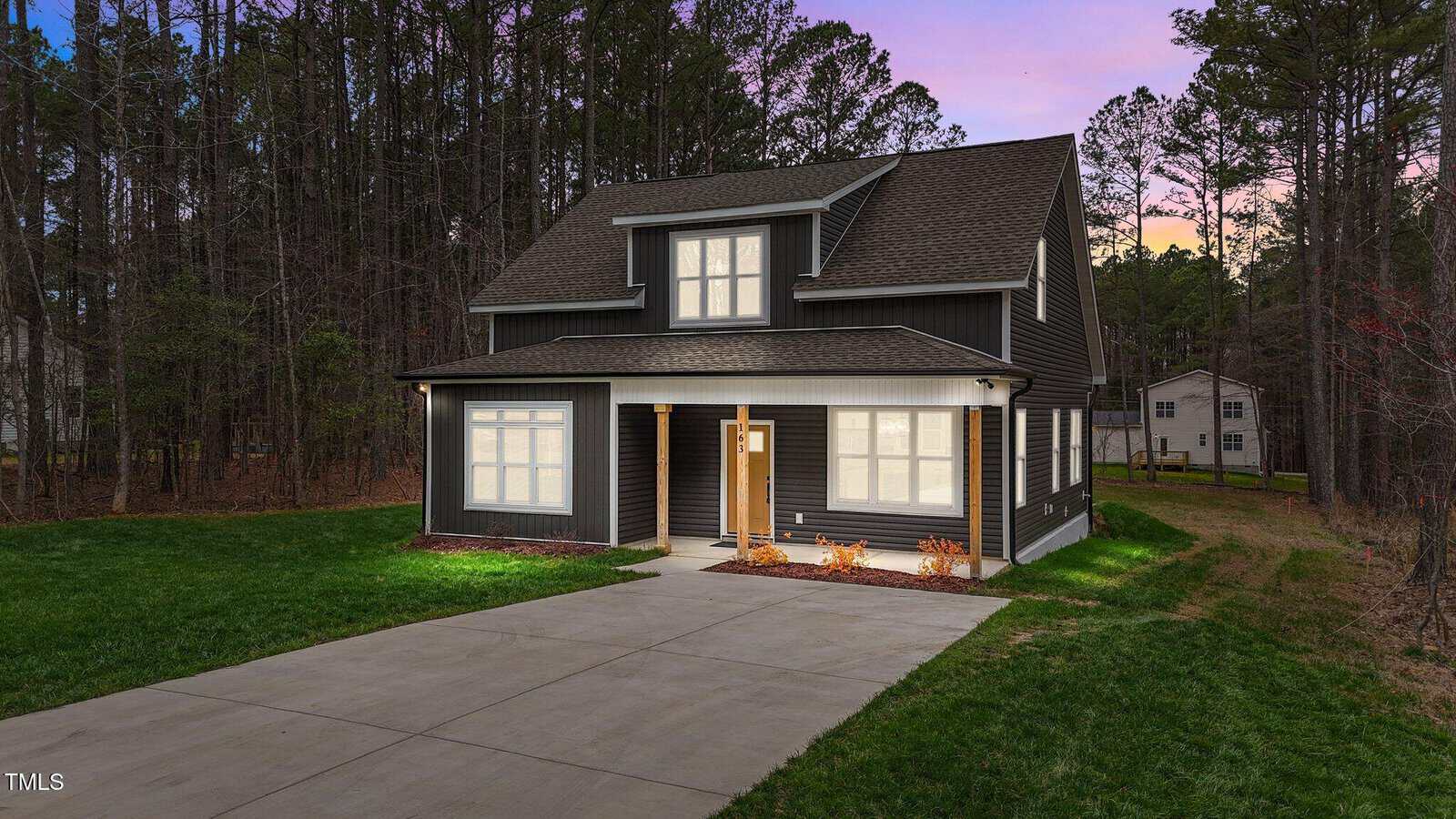 A two-story dark gray house with white trim, covered front porch, and large windows, sits beside a wooded area. A wide concrete driveway leads to the front door, and the sky shows a purple sunset.