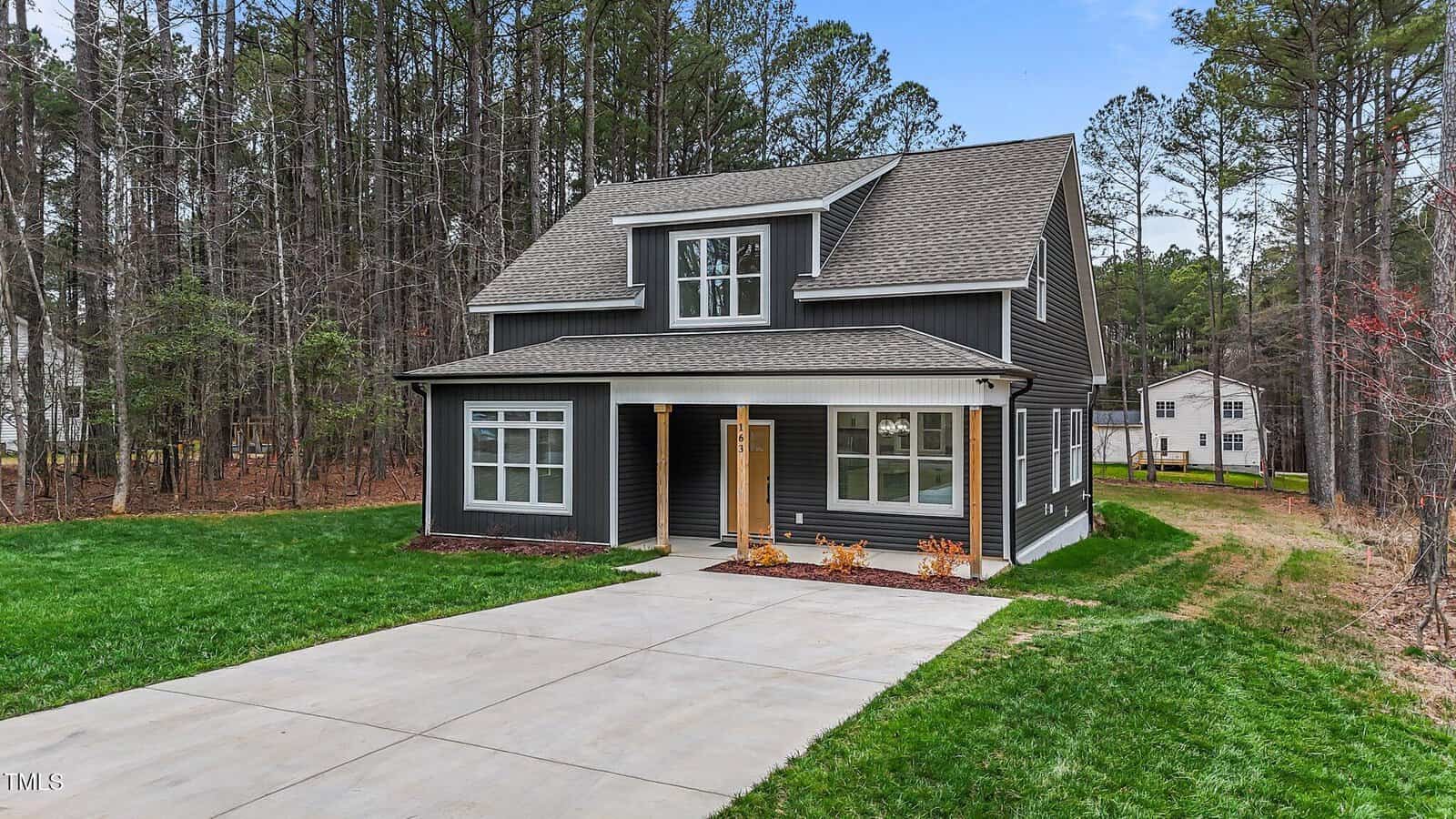 A two-story house with dark siding and white trim, a covered front porch, and large windows. The home sits next to a wooded area, with a concrete driveway and green lawn in front. Another house is visible in the background.