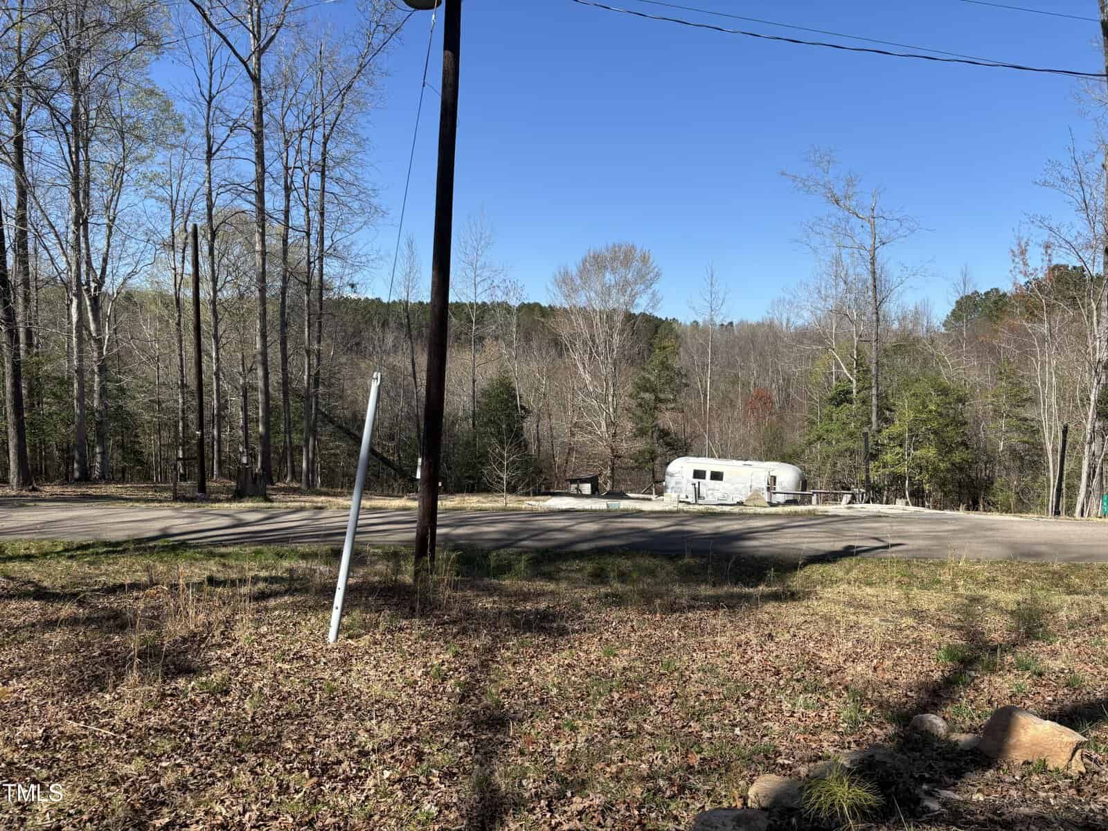 A silver trailer is parked beside a paved road surrounded by leafless and evergreen trees under a clear blue sky near 162/166 Cree Drive, Louisburg. A utility pole and a leaning road sign are visible in the foreground.